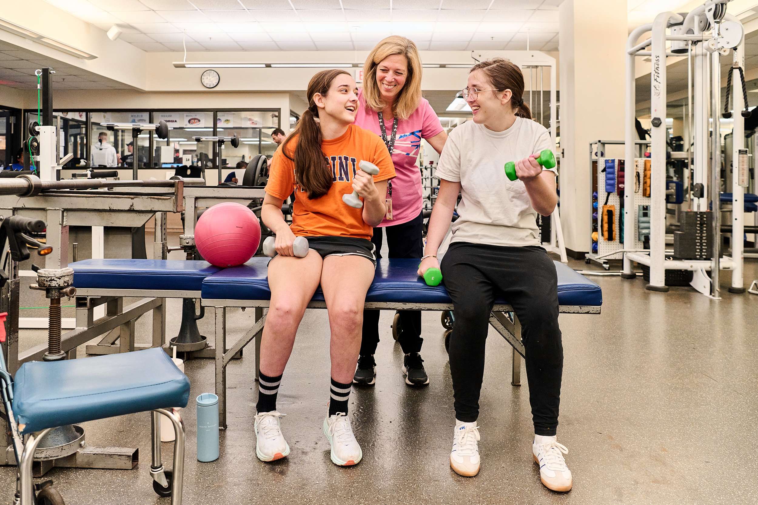 Two people are sitting on a bench in a gym, holding dumbbells and chatting with a person standing between them. Exercise equipment and a pink exercise ball are nearby. The atmosphere is friendly and focused on fitness.