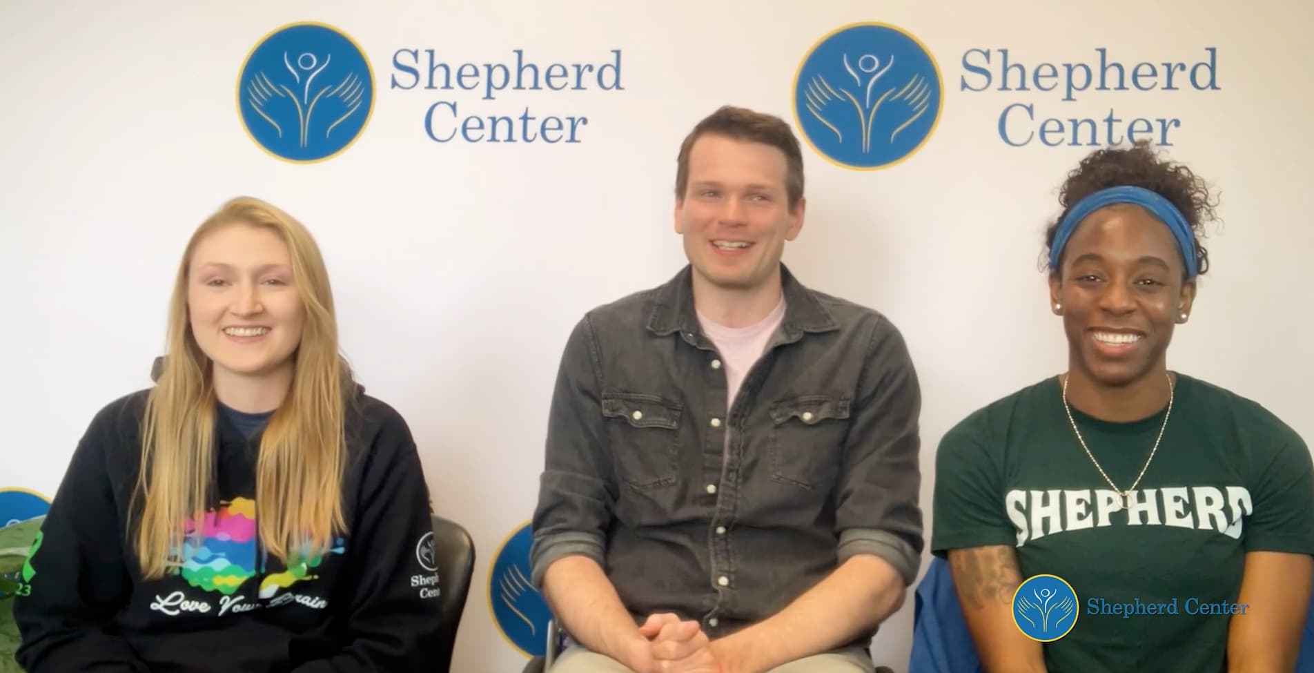 Three people are sitting side by side, smiling at the camera. Behind them is a backdrop with 