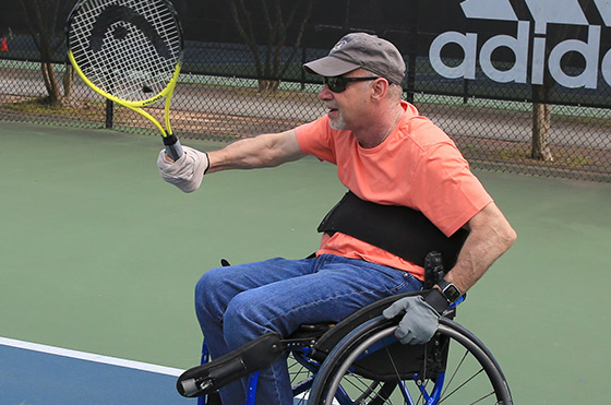 rec-therapy A man in a wheelchair, wearing a cap and sunglasses, plays tennis. He is holding a racket with a yellow frame and reaching to hit a ball. The background shows a tennis court and an Adidas banner.