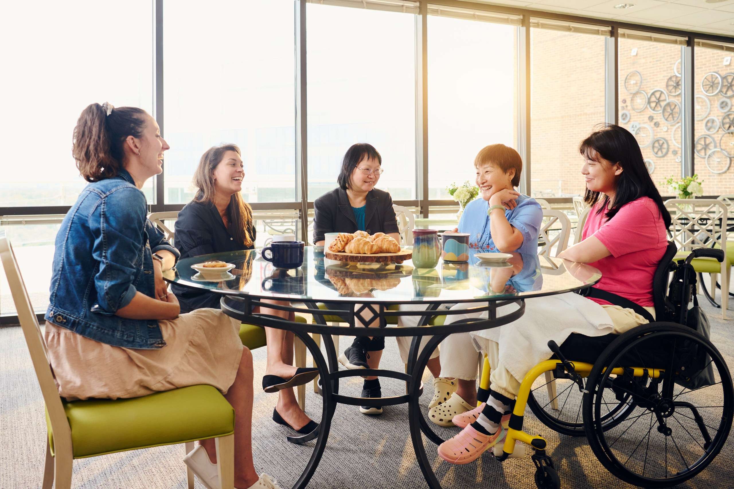 A group of five people, including a person in a wheelchair, sit around a glass table with snacks and drinks. They are engaged in a lively conversation in a bright room with large windows.