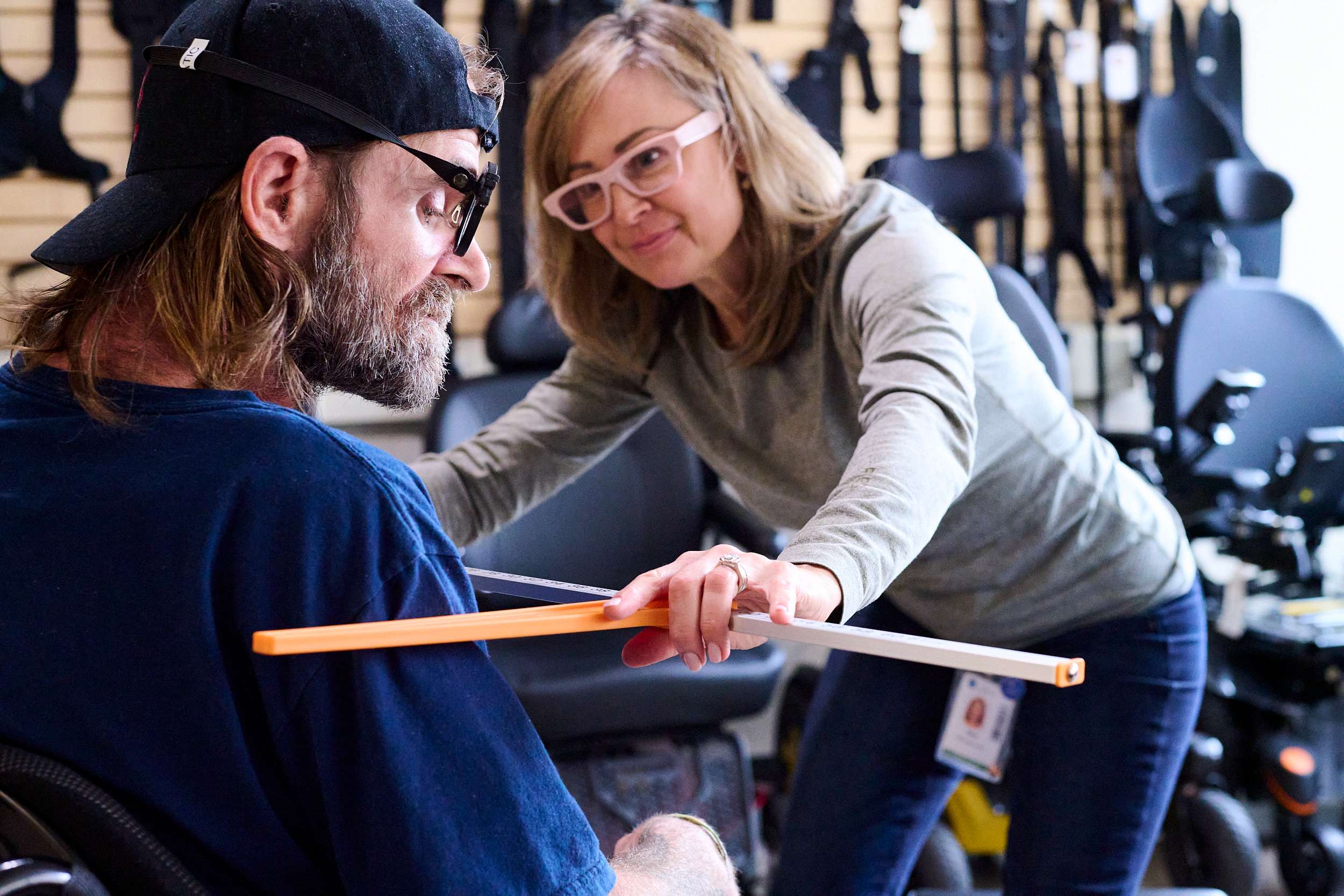 A woman assists a man in a wheelchair using a long stick for guidance in a store with mobility equipment. They appear engaged in learning or demonstration, with various assistive devices visible in the background.