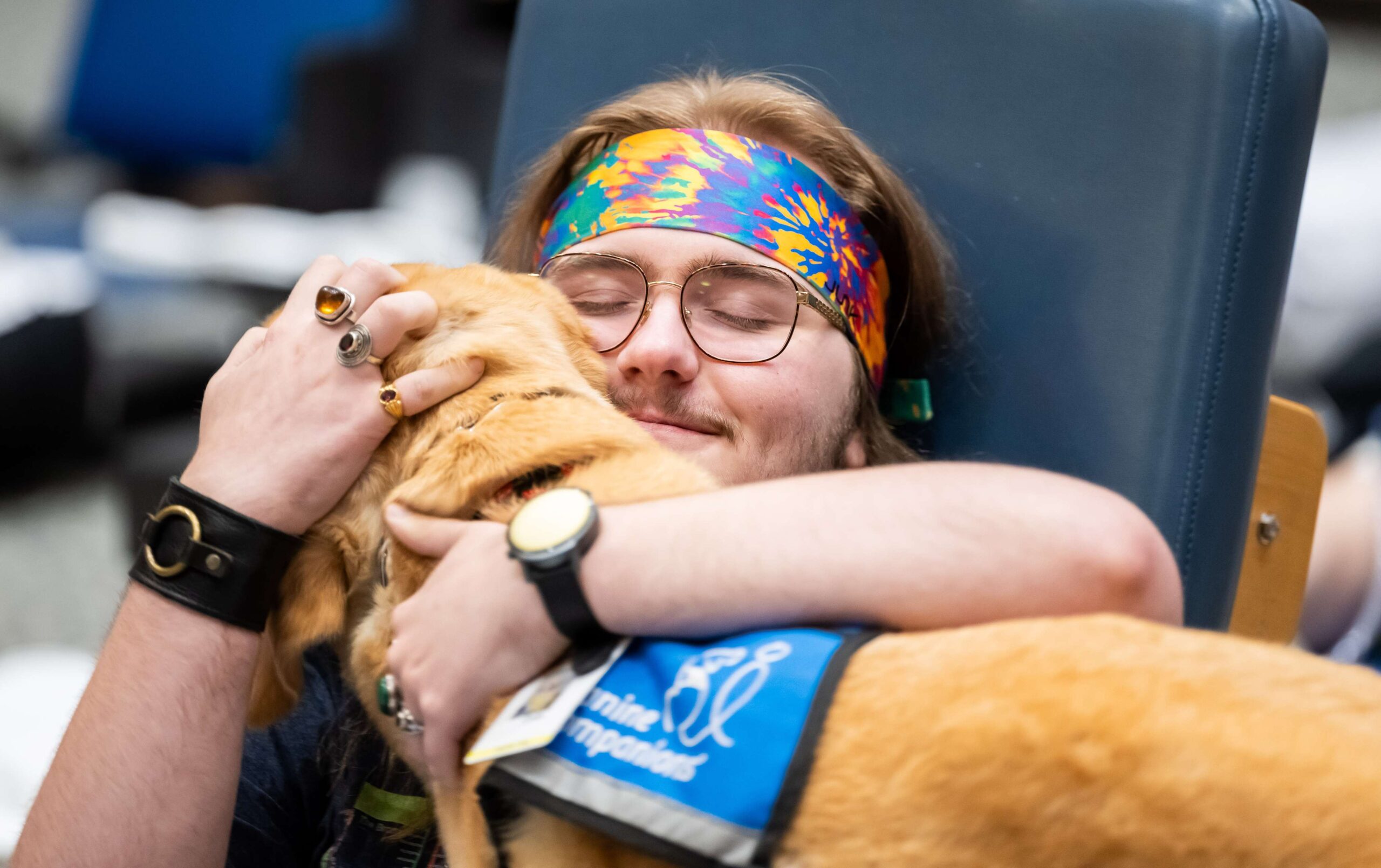 A young person with long hair and a colorful headband joyfully hugs a golden retriever wearing a blue vest. The person is seated and wearing glasses and rings, with a content smile on their face.