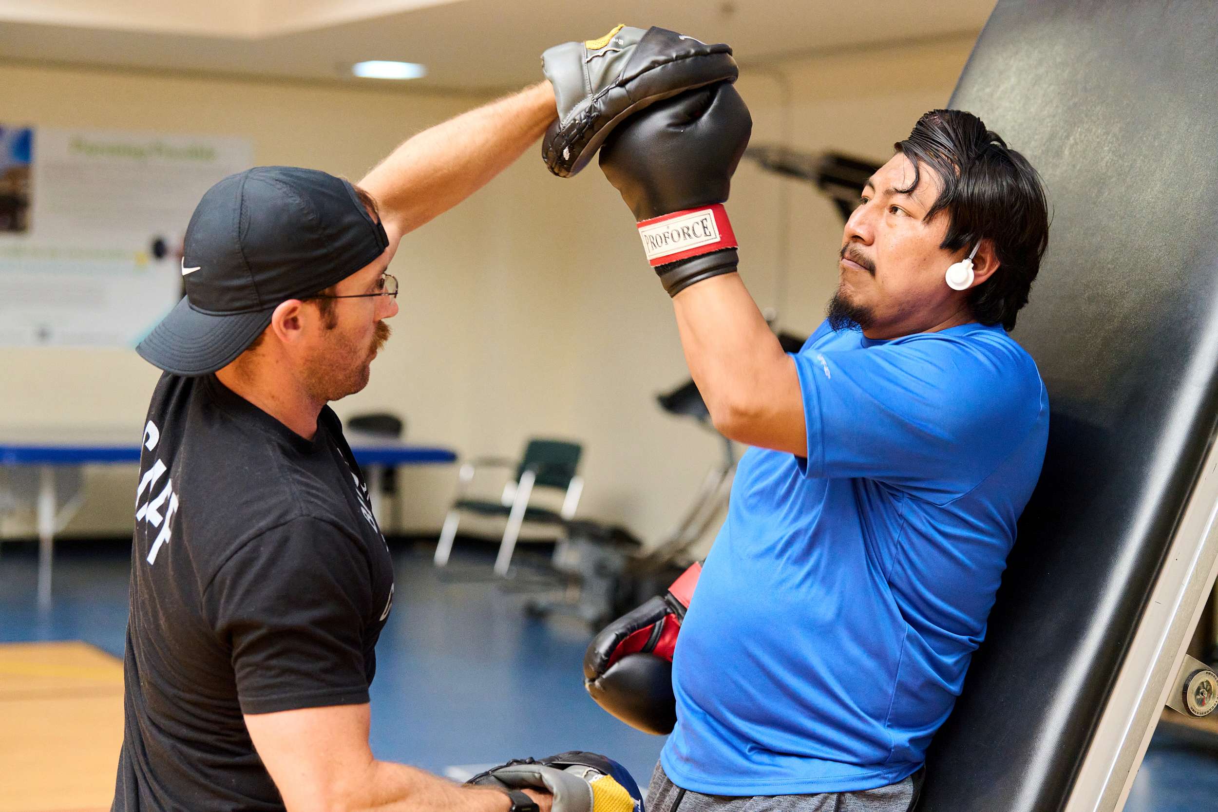 A man in a blue shirt practices boxing with a trainer in a black cap holding focus mitts. They are in a gym environment, and the man is leaning back slightly, wearing earphones.