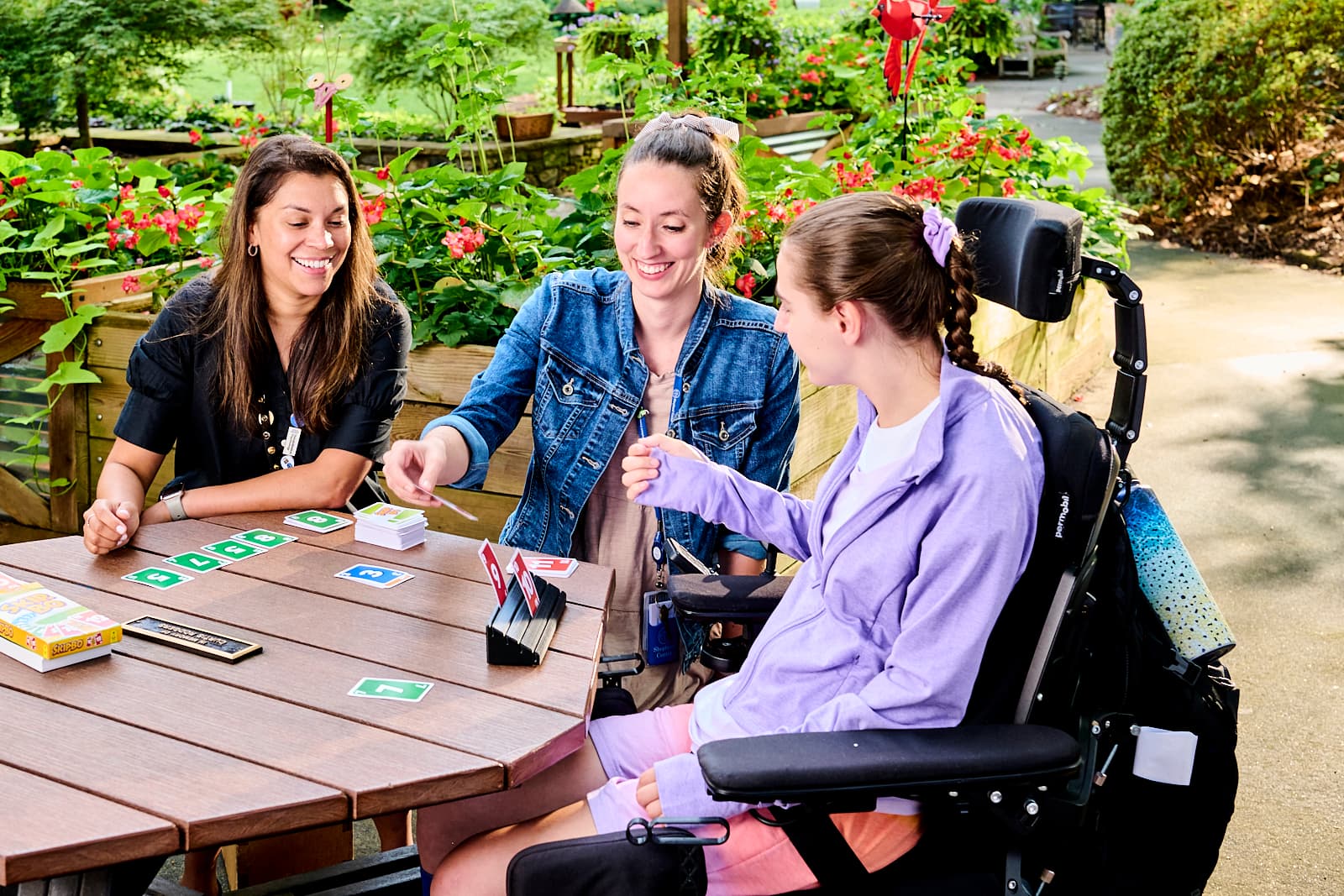 Three people are playing a card game at an outdoor table surrounded by lush greenery and flowers. One person is using a wheelchair, and they are all smiling and engaged in the game.