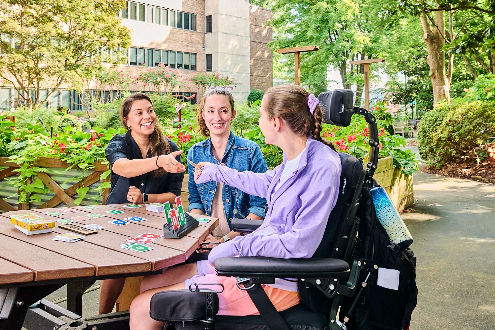 Three women are sitting at a round outdoor table playing a card game. One woman is in a wheelchair, and all are smiling and engaged. The setting is a garden with green trees and bushes, and a building is in the background.