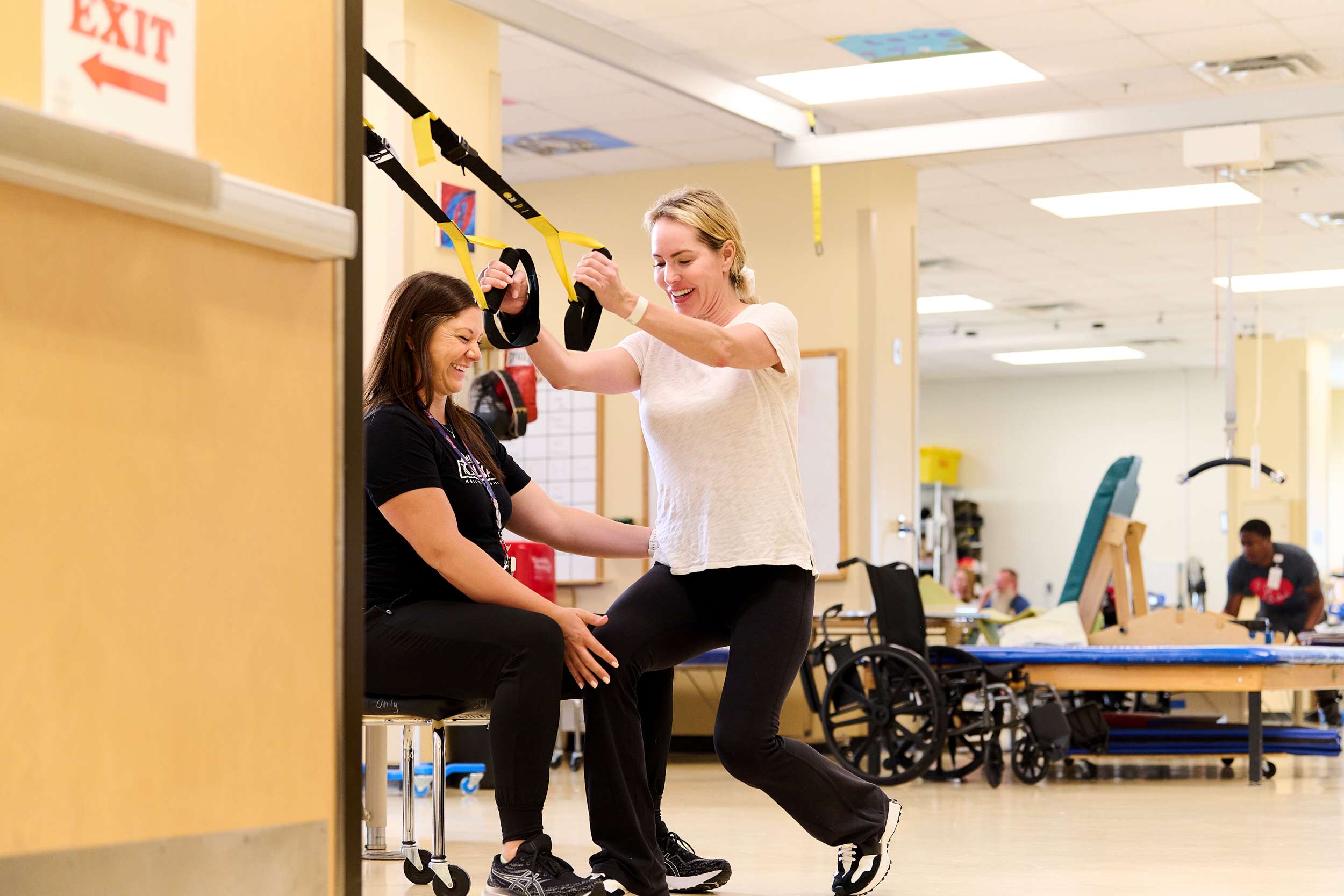 A woman assists another woman using TRX suspension straps in a physical therapy setting. The woman using the straps is smiling, while the assistant supports her from a seated position. Exercise equipment and a wheelchair are visible in the background.