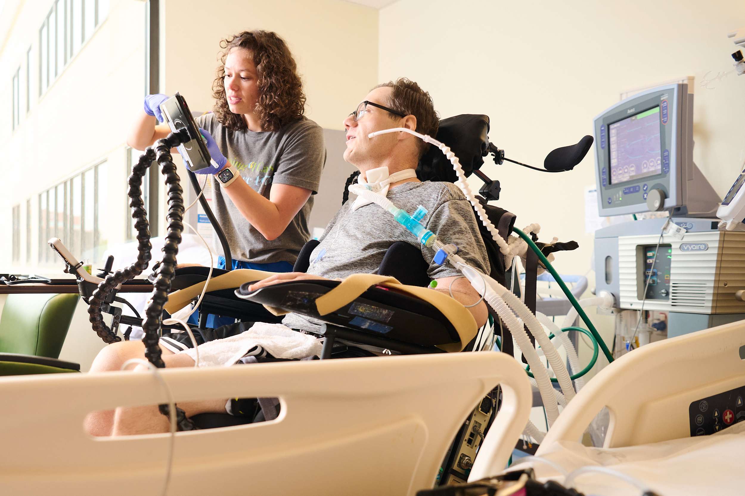 A healthcare worker assists a patient in a hospital room. The patient is seated in a specialized wheelchair connected to medical equipment. The worker adjusts a device, while various monitors are visible in the background.