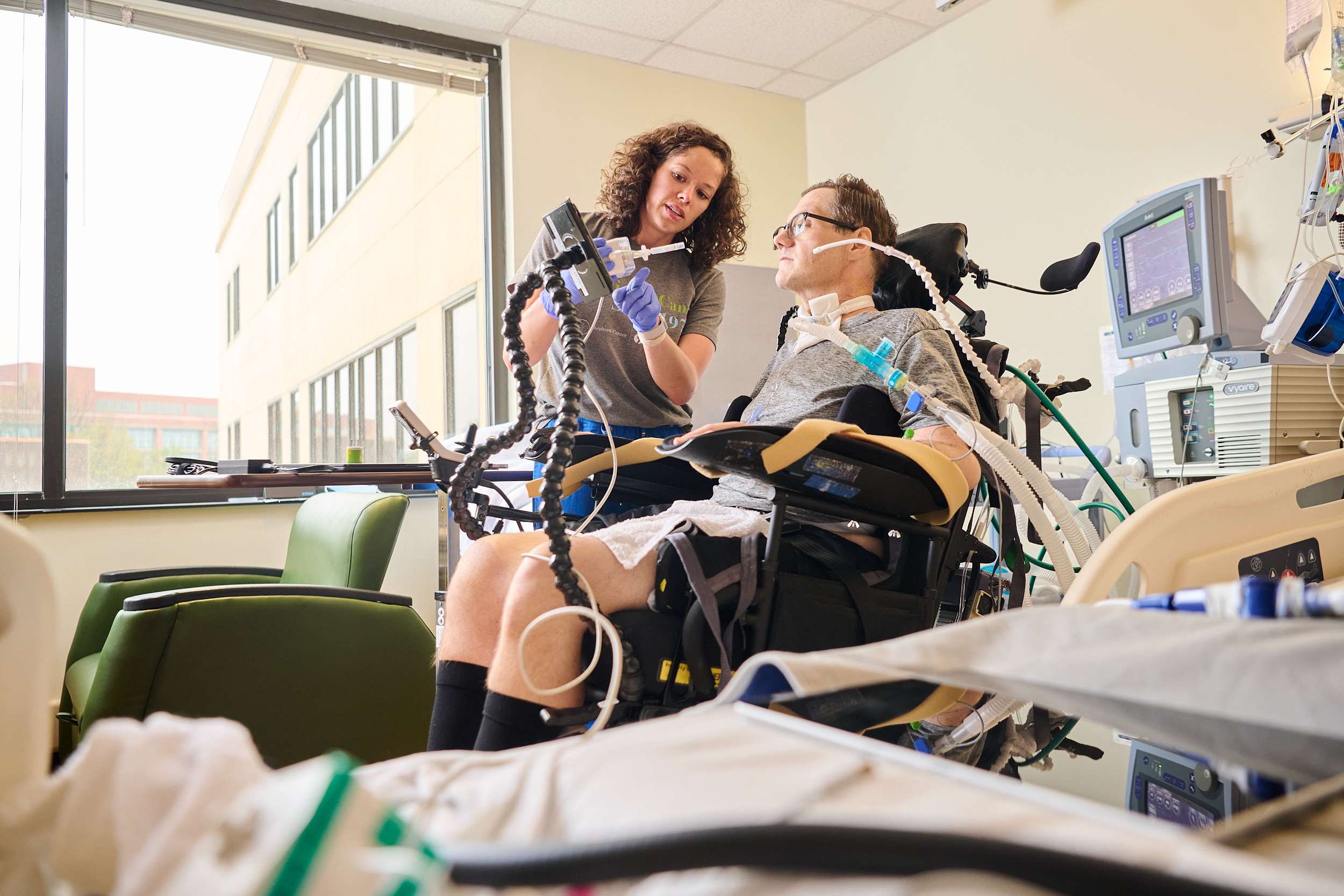 A person in a wheelchair is in a hospital room, connected to medical equipment. A assistive technology therapist stands beside them, holding a device and engaging with the patient.