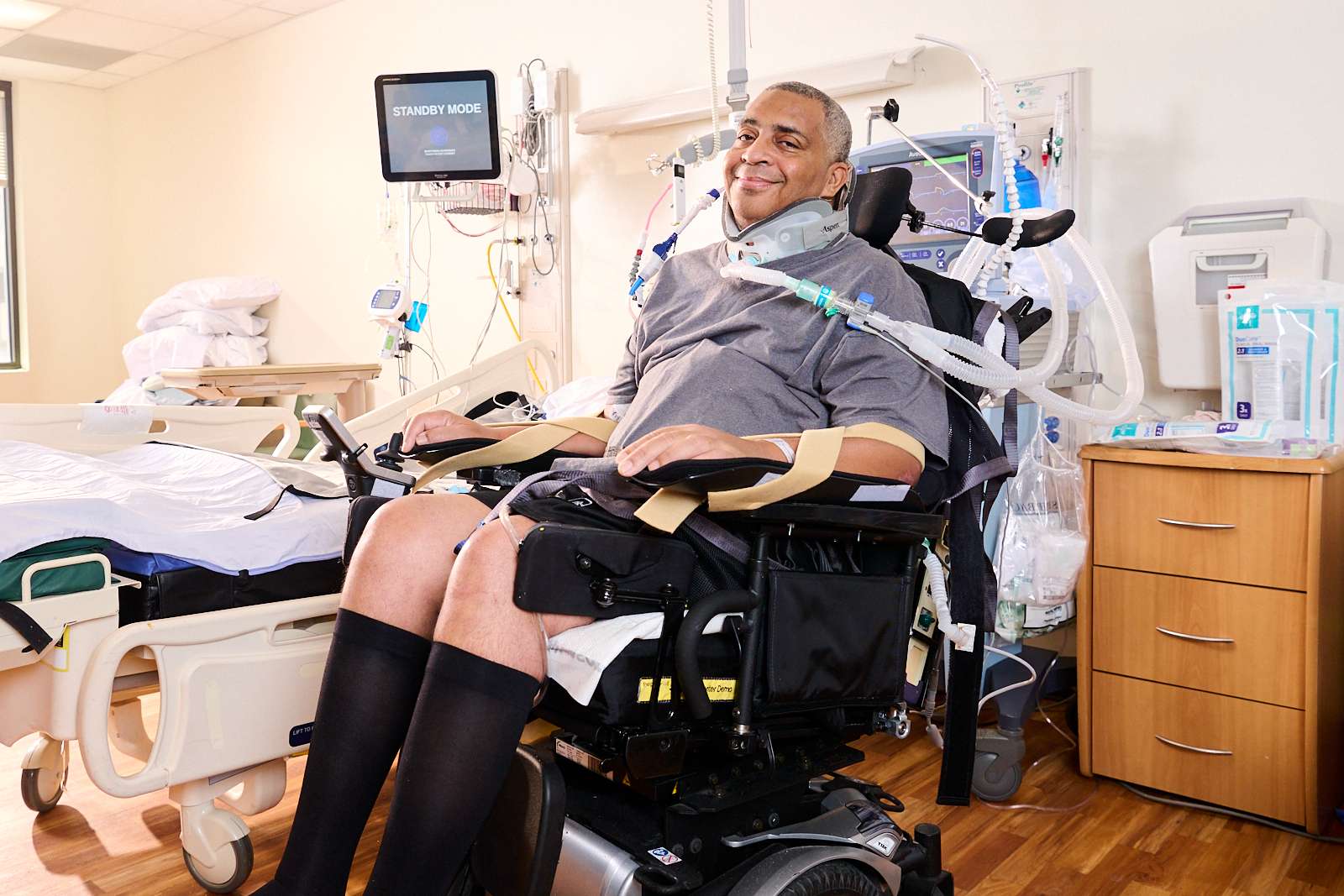 A person in a motorized wheelchair smiles in a hospital room. The room contains medical equipment, a bed, and a monitor. The person is wearing a gray shirt and black knee-high socks and has a ventilator tube attached.