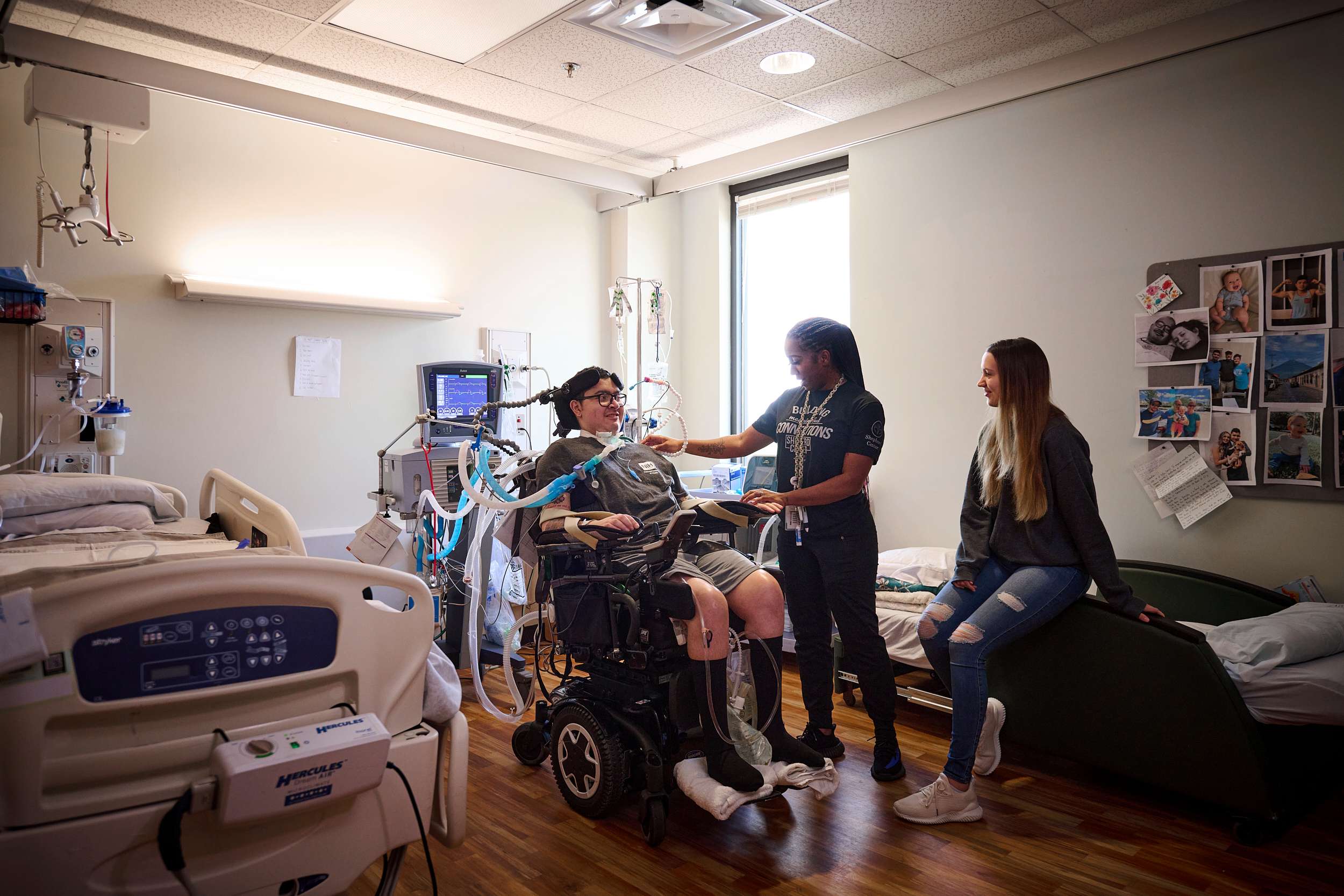 A person in a wheelchair is in a hospital room, connected to medical equipment. Two people are with them; one stands beside, touching the wheelchair user, while the other sits on a nearby bed. The room is filled with medical devices and personal photos on the wall.