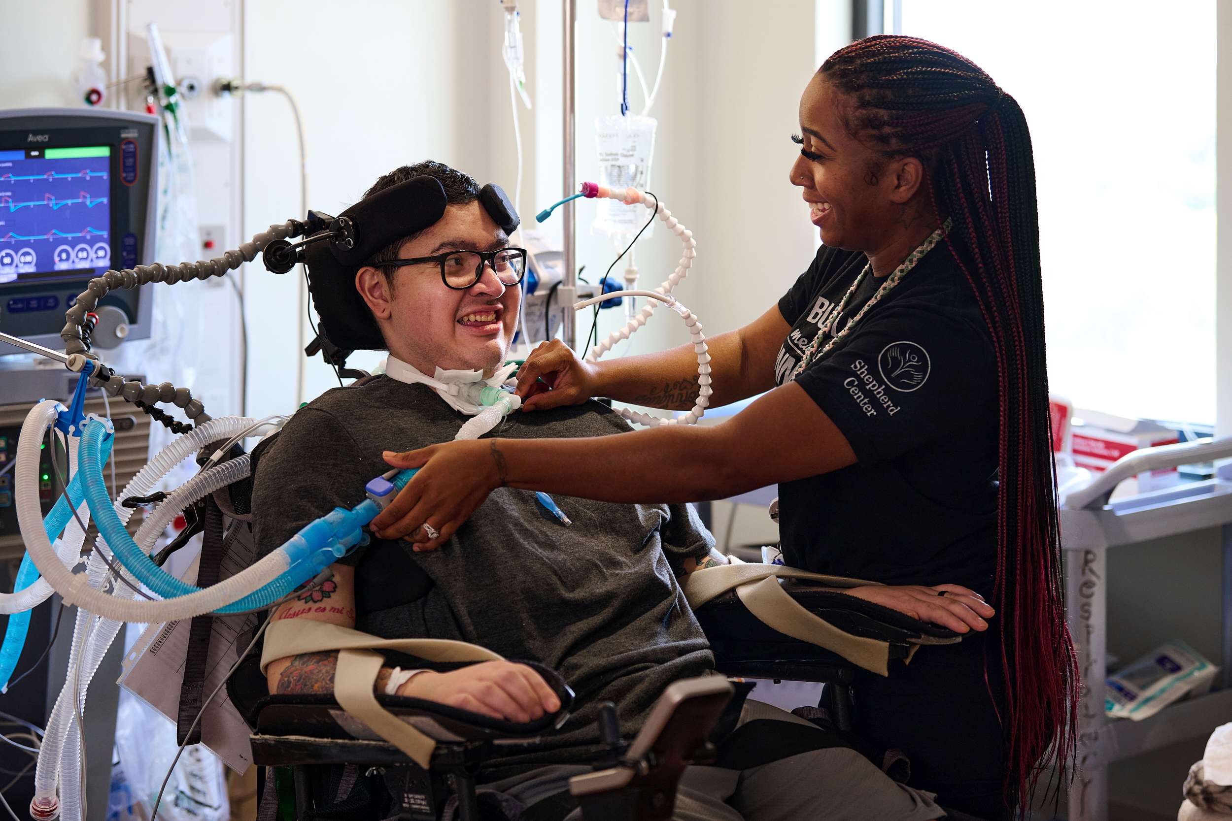 A patient in a wheelchair with medical equipment smiles as a caregiver adjusts a device on them. The caregiver is wearing a black shirt and smiling, creating a positive atmosphere in a hospital setting.