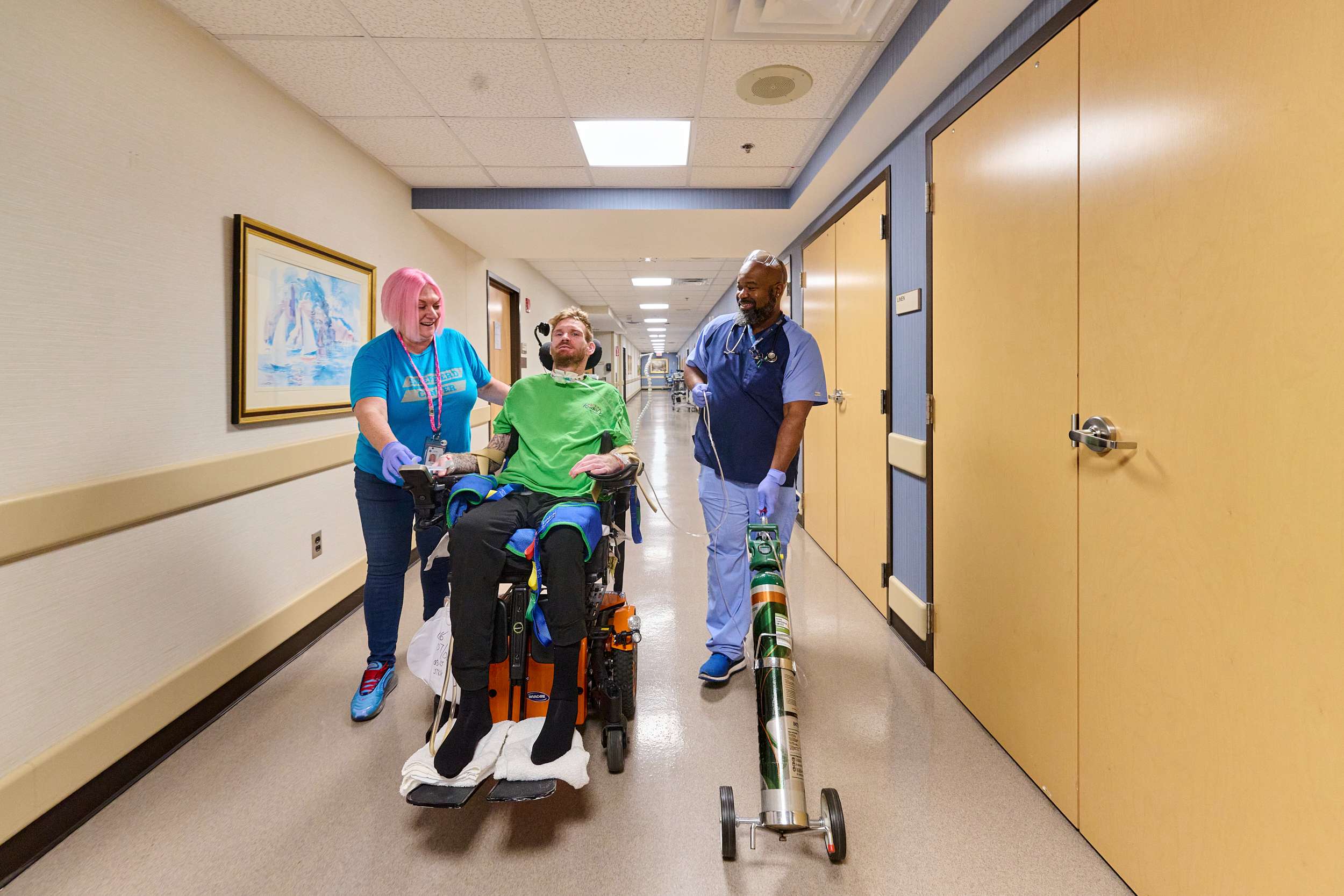 A person in a green shirt is sitting in a wheelchair, accompanied by two respiratory therapists in a hospital hallway. One therapist pulls an oxygen tank cart. They are smiling and appear to be engaged in conversation.