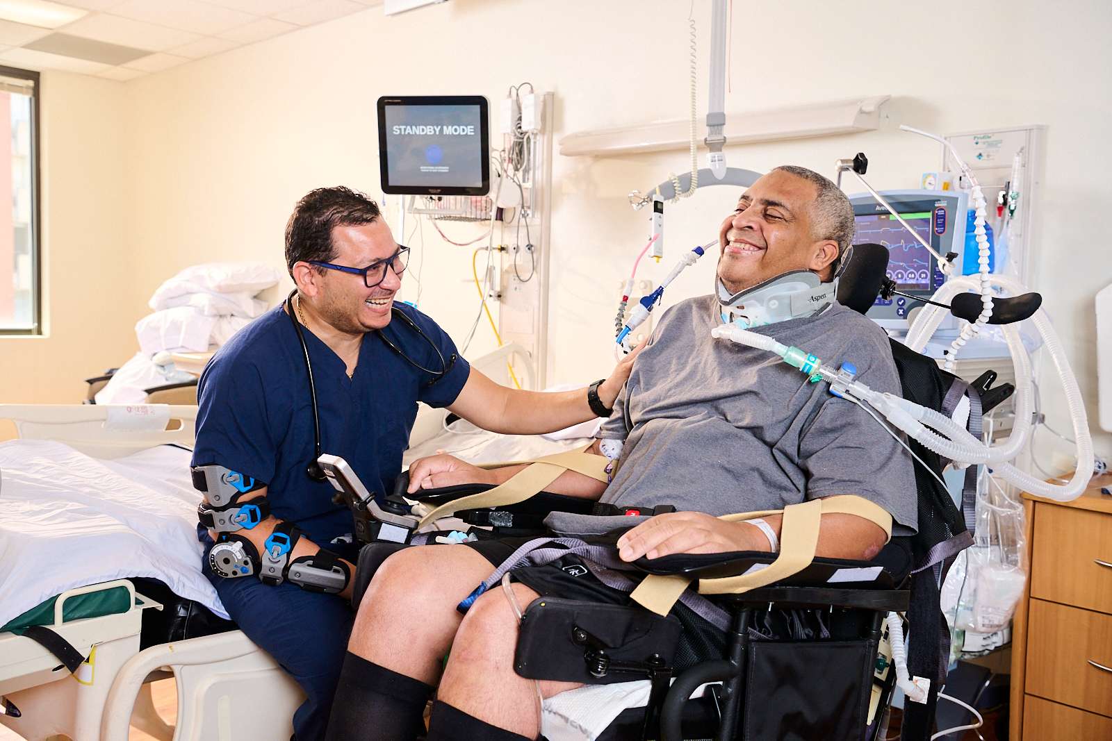 A healthcare professional in scrubs sits beside a smiling patient in a hospital room. The patient, wearing a neck brace and connected to medical equipment, appears to be in a wheelchair. The room is filled with medical devices and bedding.