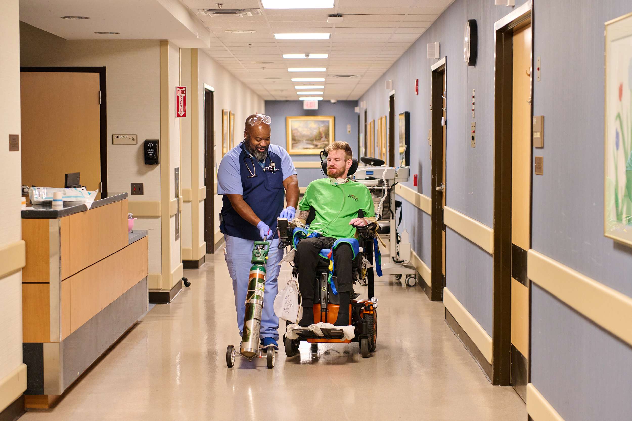 A nurse walks alongside a man in a motorized wheelchair down a hospital corridor. The man in the wheelchair wears a green jacket and appears to be in conversation with the nurse, who is wearing blue scrubs. The hallway is brightly lit and decorated with artwork.