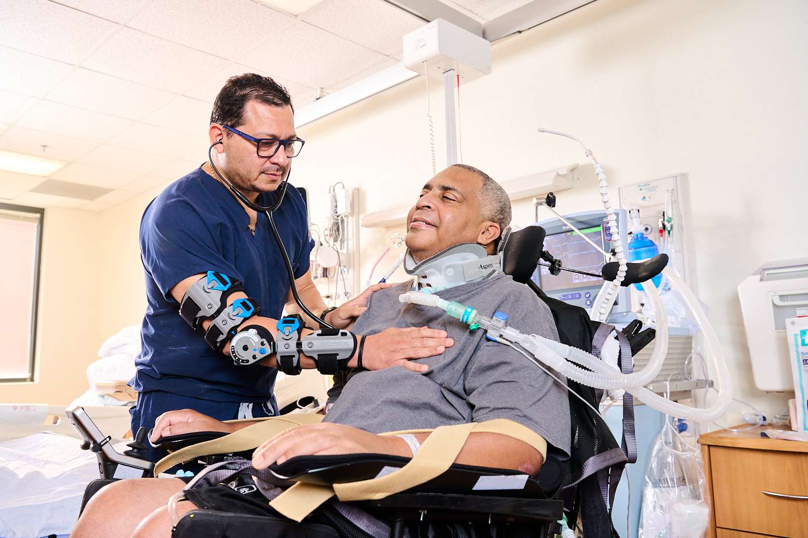 A pulmonologist in blue scrubs uses a stethoscope to listen to the heartbeat of a patient in a wheelchair wearing a neck brace and connected to a ventilator.