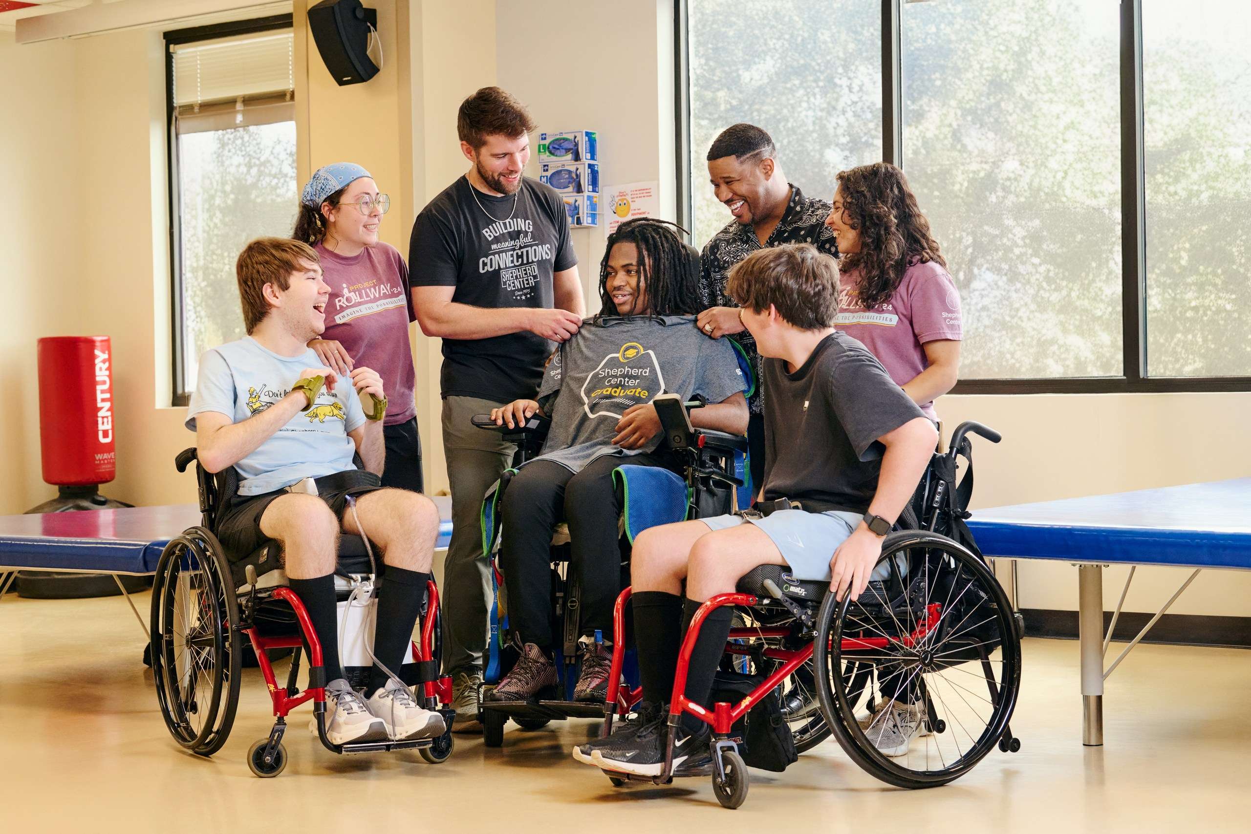 A diverse group of seven people, including five in wheelchairs, gather in a bright room. They’re smiling, and one person receives a t-shirt. A large window and exercise equipment are visible in the background.