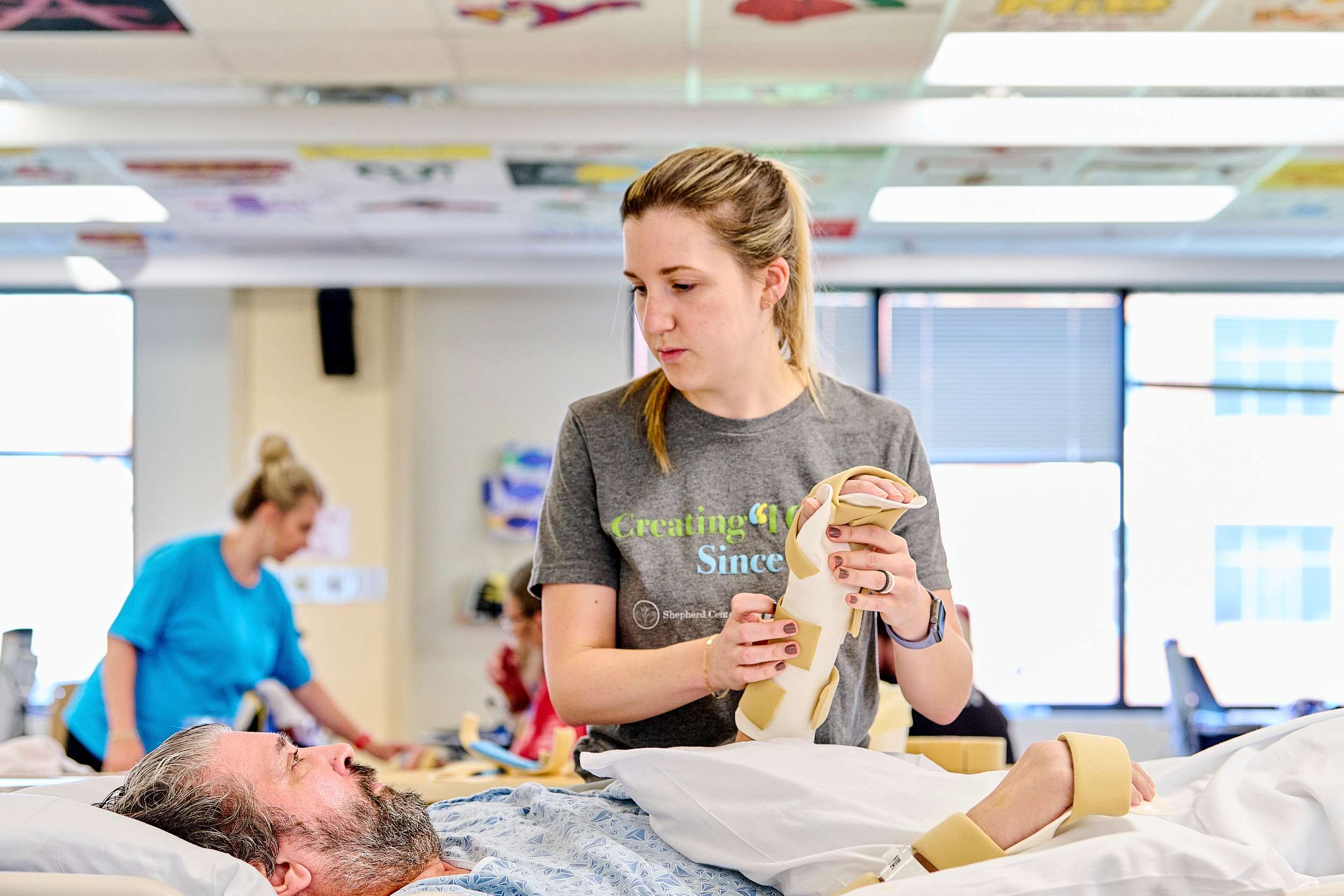 A healthcare worker assists an older man in a hospital bed, fitting his foot with a medical brace. Another staff member is visible in the background. The room is brightly lit with windows and ceiling decorations.