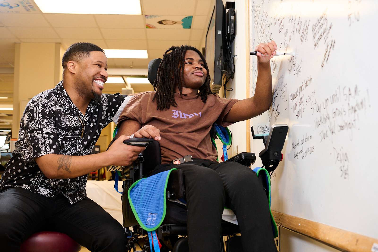 A man in a black and white shirt smiles and supports another man in a wheelchair who is writing on a whiteboard. They appear to be collaborating in a brightly lit room.