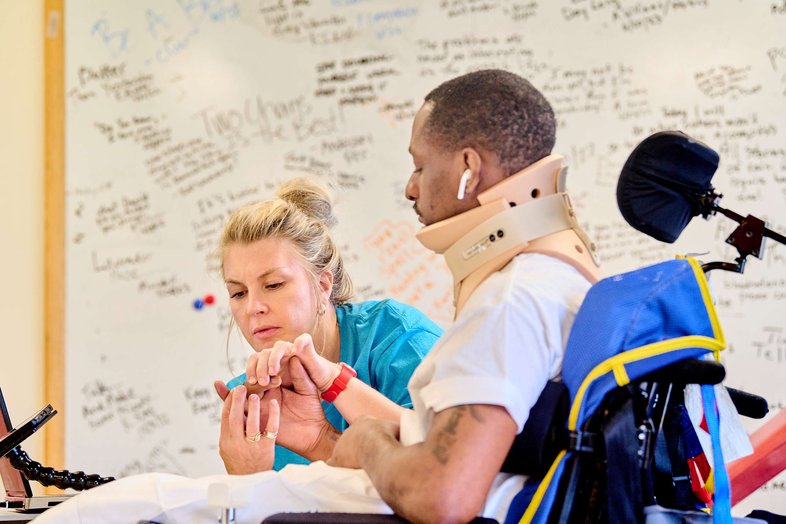 A healthcare worker assists a person in a wheelchair wearing a neck brace. They are in a room with a large whiteboard covered in writing. The healthcare worker is checking the person's hand.