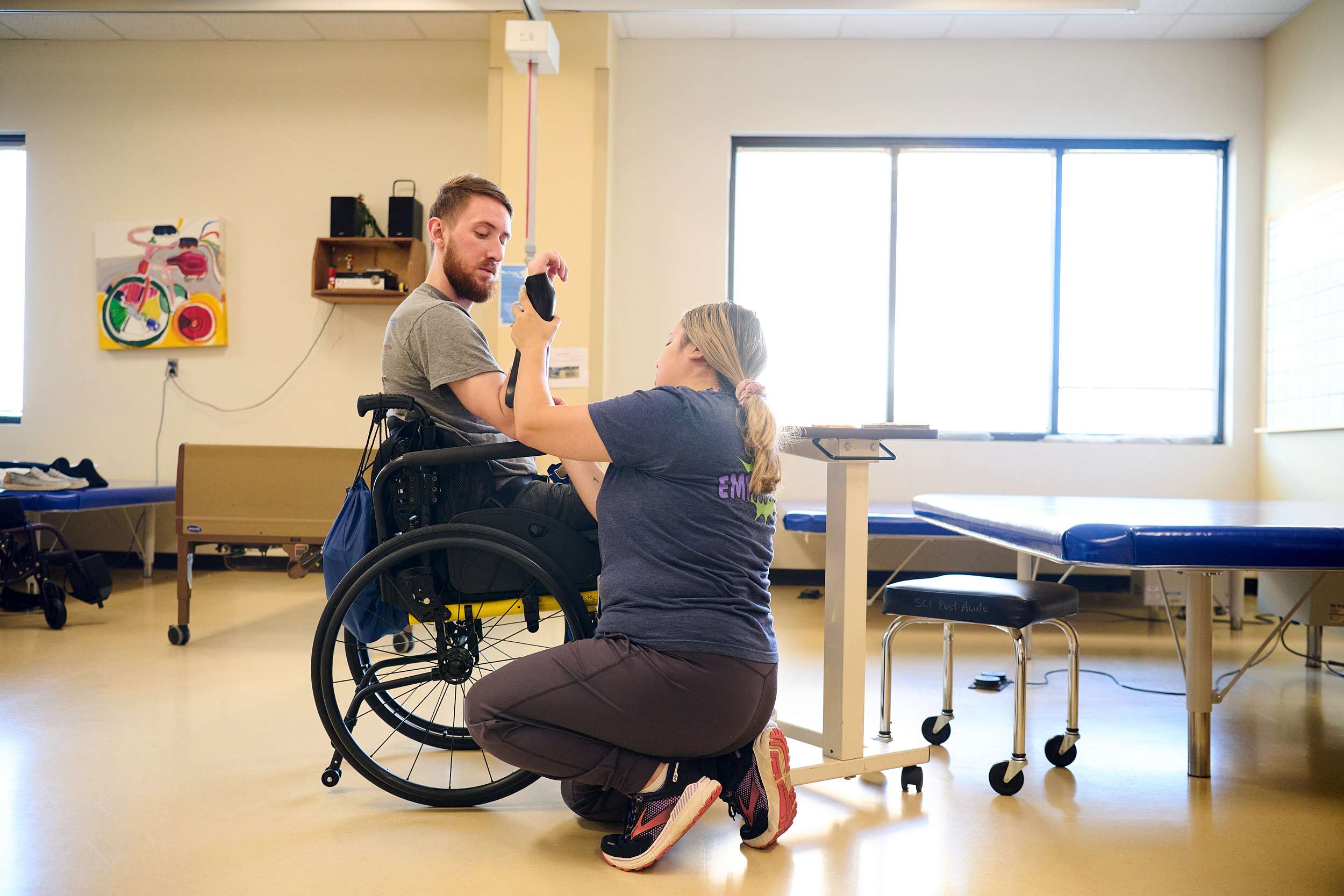 A physical therapist helps a person in a wheelchair with a stretching exercise in a therapy room. Sunlight streams through large windows, illuminating the space filled with medical equipment and colorful artwork on the walls.