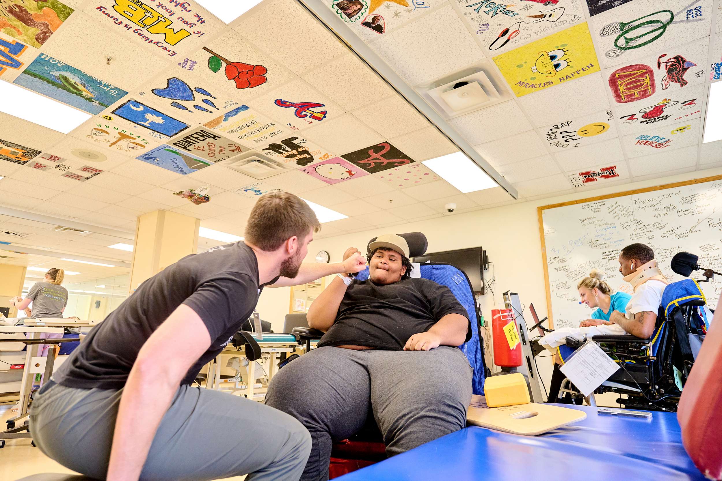 A physical therapist interacts with a patient seated in a hospital room. The ceiling is decorated with colorful art tiles. Other patients and therapists are seen in the background engaging in various activities.