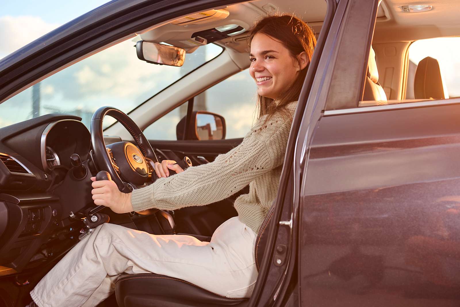 A person with long hair sits confidently in the driver's seat of a car, smiling. The car door is open, and the sun sets in the background, casting a warm glow inside the vehicle.