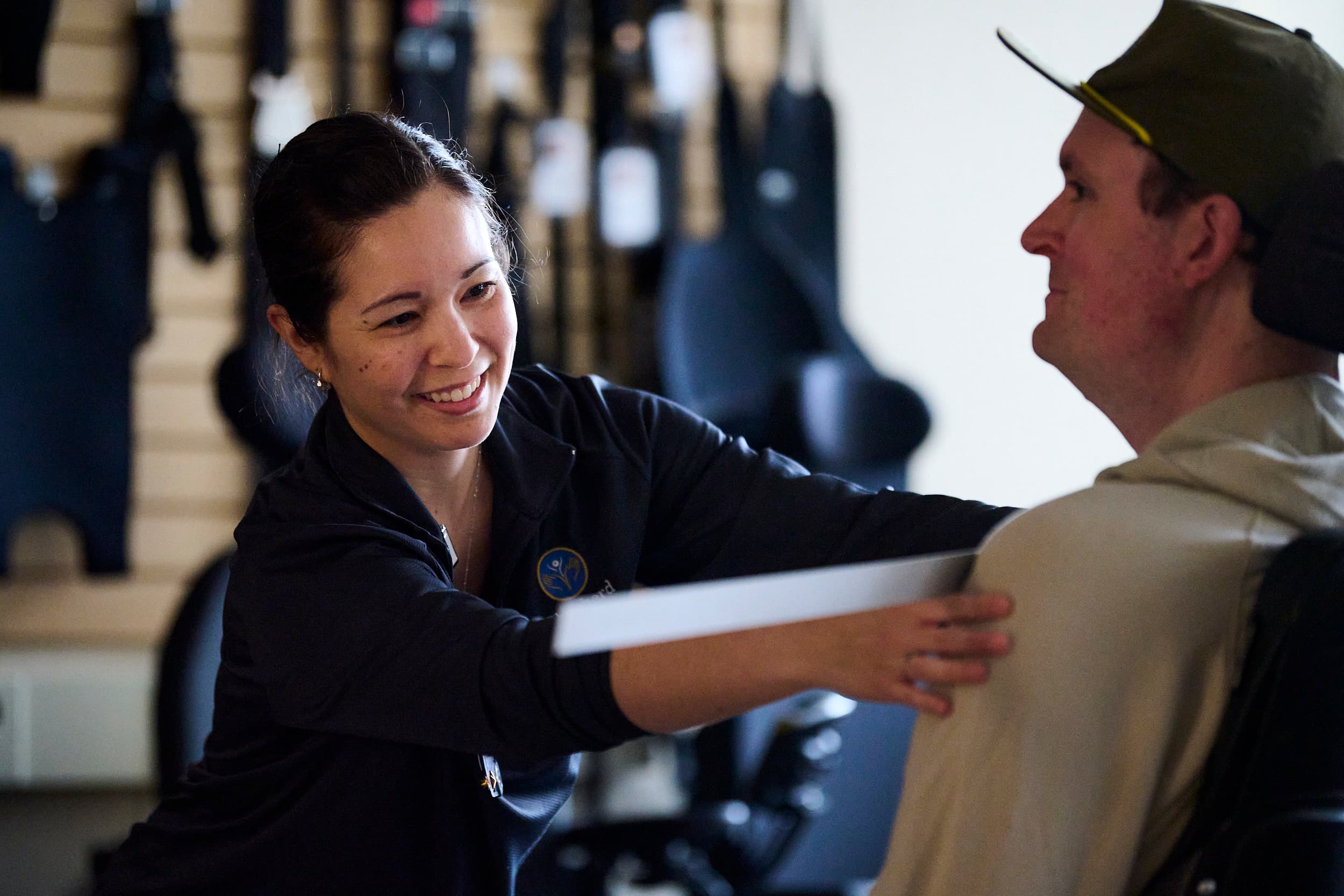 A woman measures a man's shoulder using a ruler. The man is seated in a wheelchair, and they are in a room with various equipment on the wall. The woman is smiling as she works.