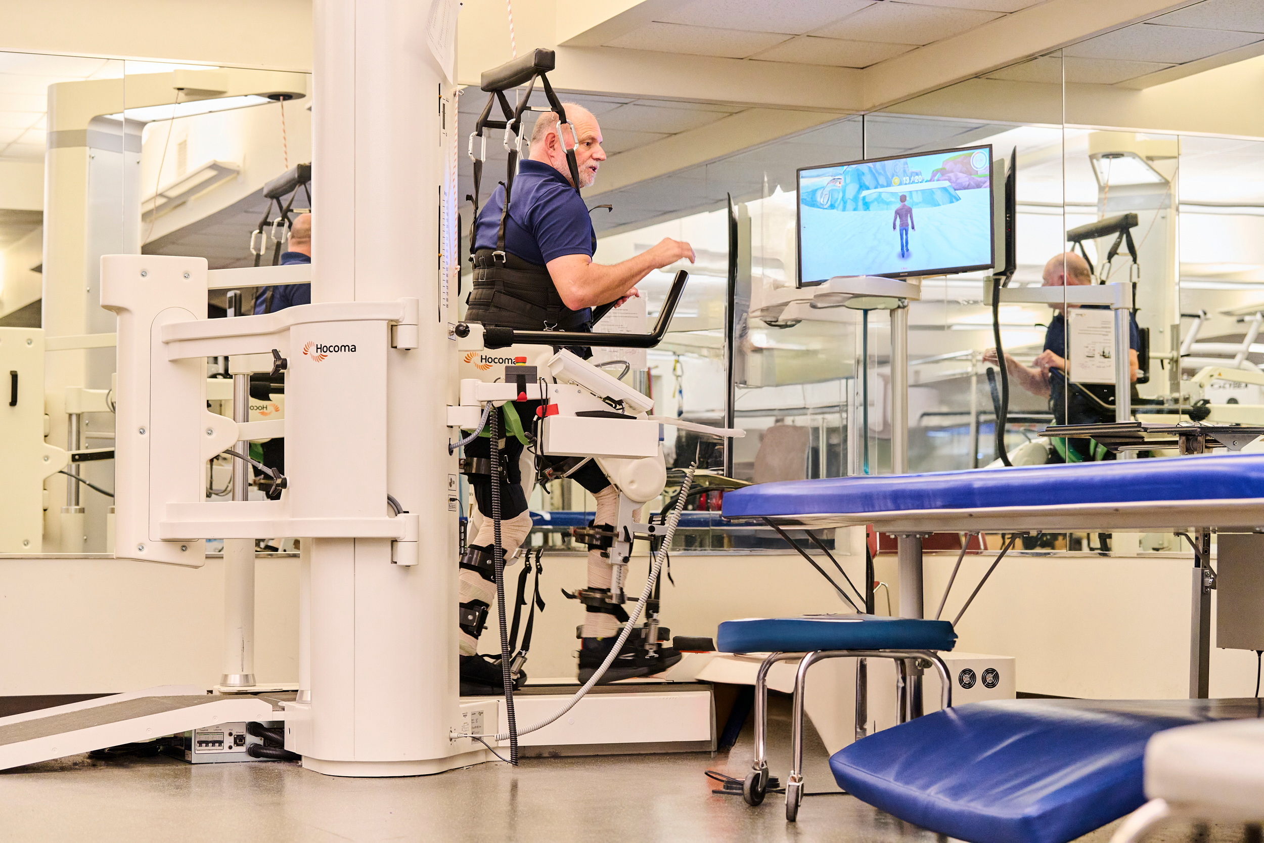 A man undergoing therapy with the help of a robotic exoskeleton and a therapist at a rehabilitation center. The therapist wears a jacket with 