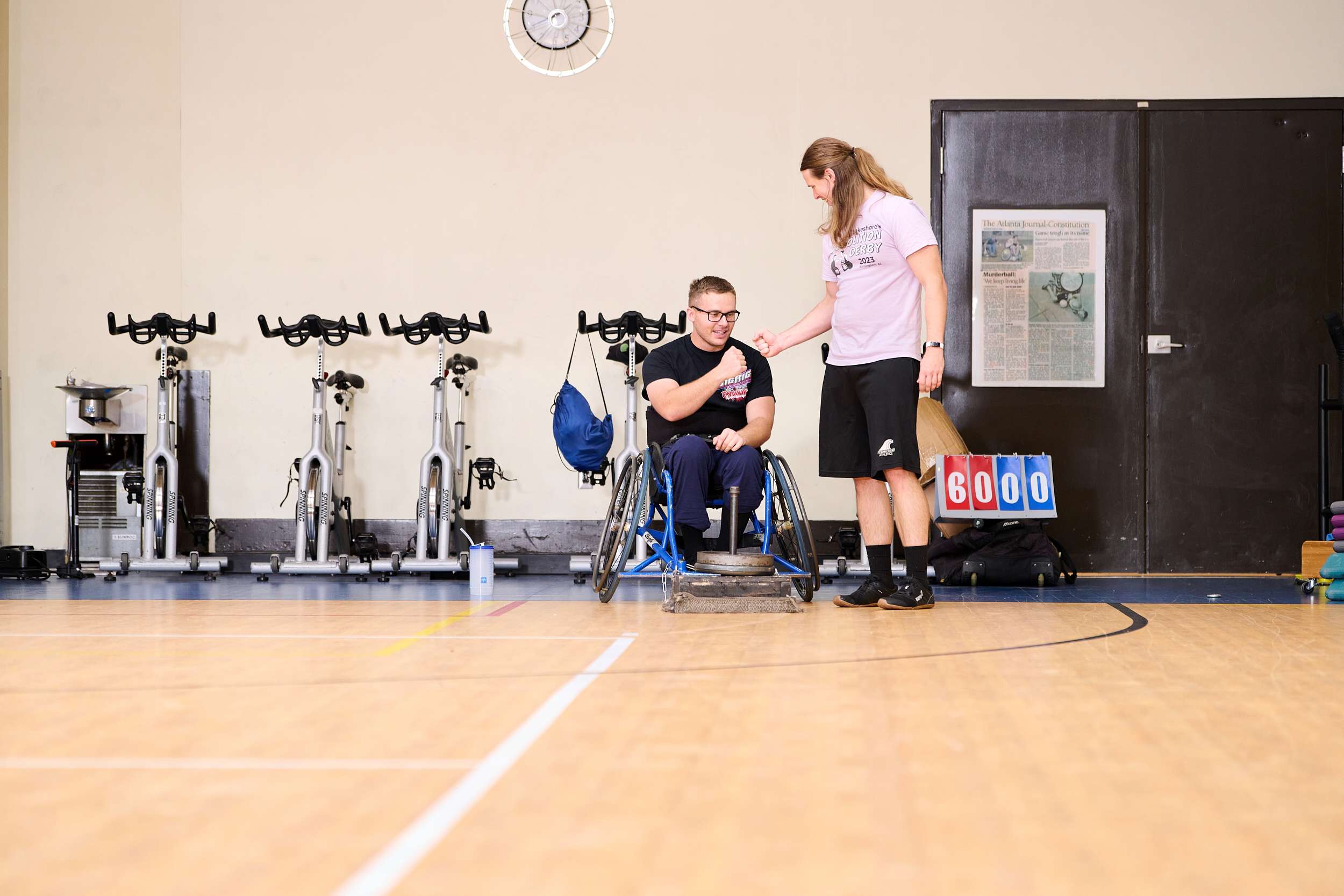 A person in a wheelchair fist bumps another person standing beside them in a gym. They are on a basketball court, with stationary bikes and a scoreboard in the background.