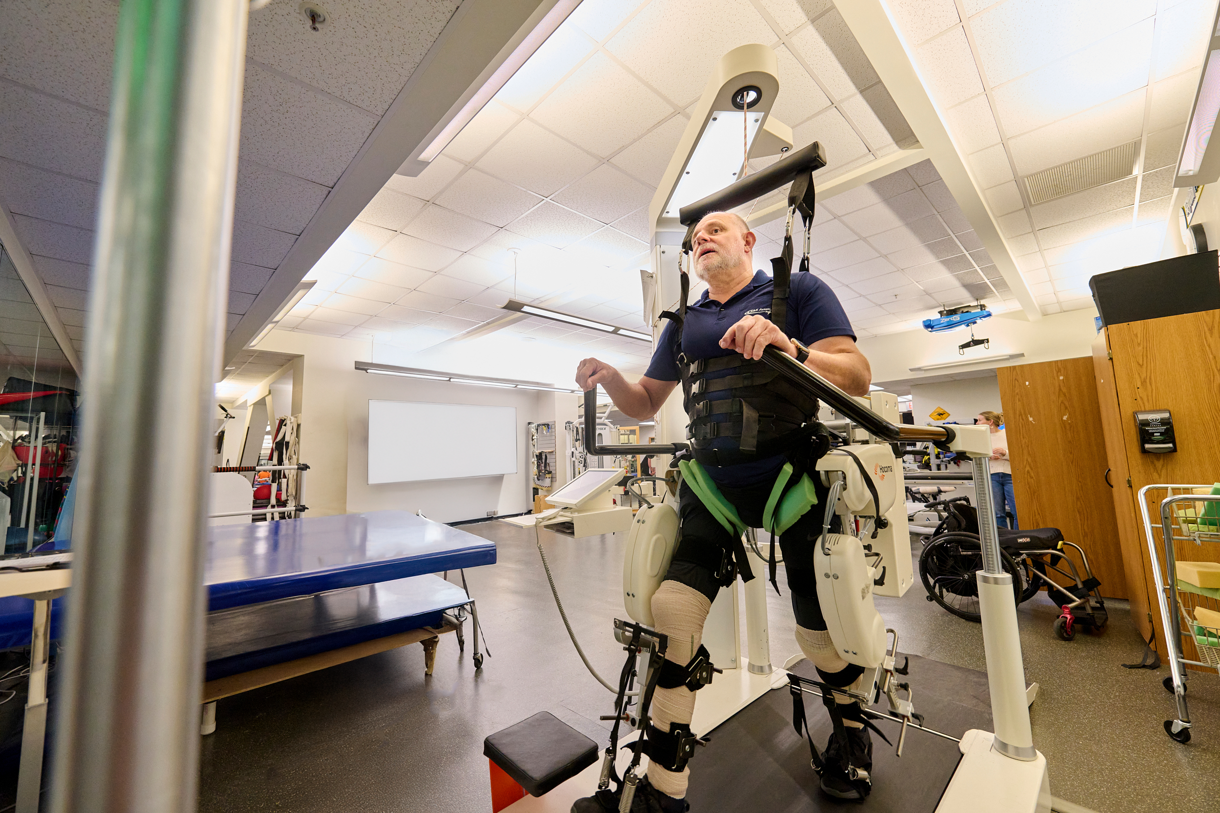 A man is standing on a treadmill-like device wearing an exoskeleton. Another man is nearby, observing and assisting. The setting appears to be a rehabilitation facility with various exercise equipment in the background.