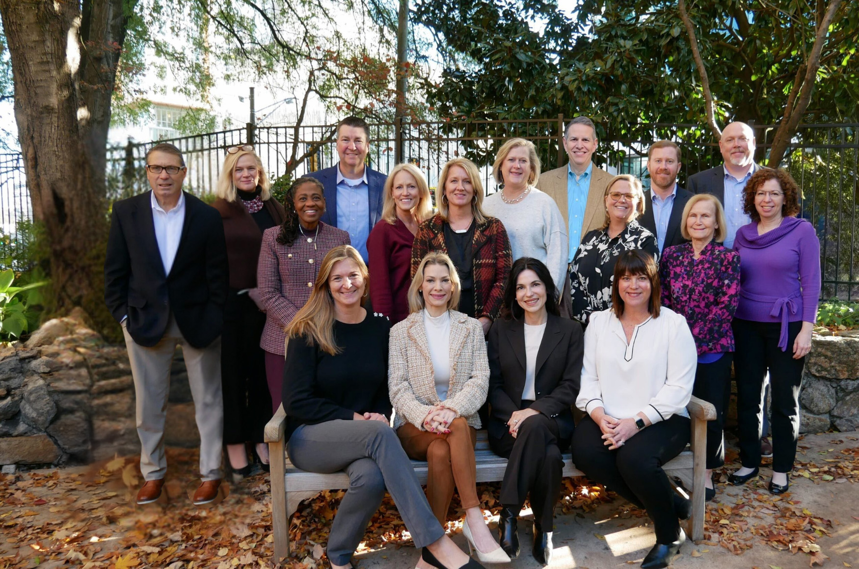 A group of 18 people is gathered outdoors, posing for a photo. They are positioned in rows, with three people seated on a bench in the front. Fallen leaves and trees in the background suggest an autumn setting. Everyone is dressed in business casual attire.