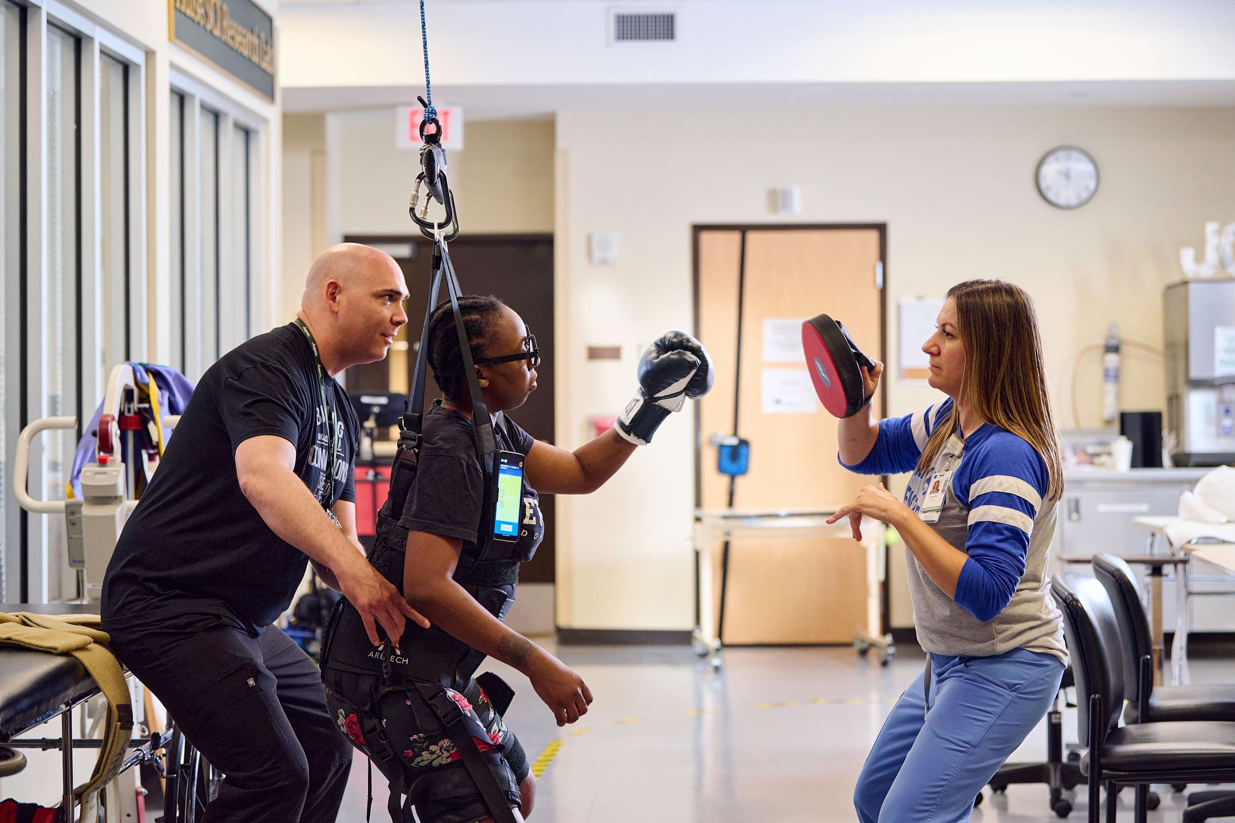 A woman wearing assistive gear practices punching a pad held by a trainer. Another person provides support from behind. The setting appears to be a rehabilitation or physical therapy center with various equipment in the background.