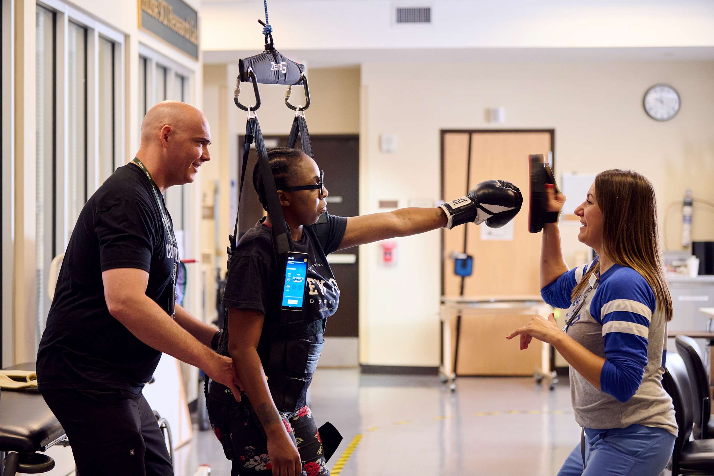 A woman in an exoskeleton suit is practicing boxing with a trainer, who holds pads. Another individual supports her. They are in a rehabilitation facility, with equipment visible in the background.