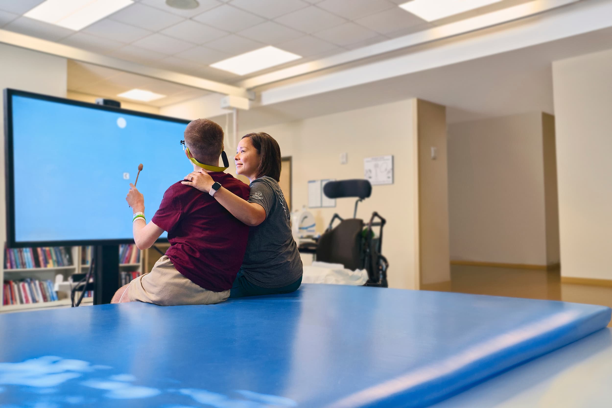 A man and a woman sit on a large blue mat in a rehabilitation center, facing a large screen. The man is holding a wand, interacting with the screen. Shelves with books are in the background, and a wheelchair is nearby.