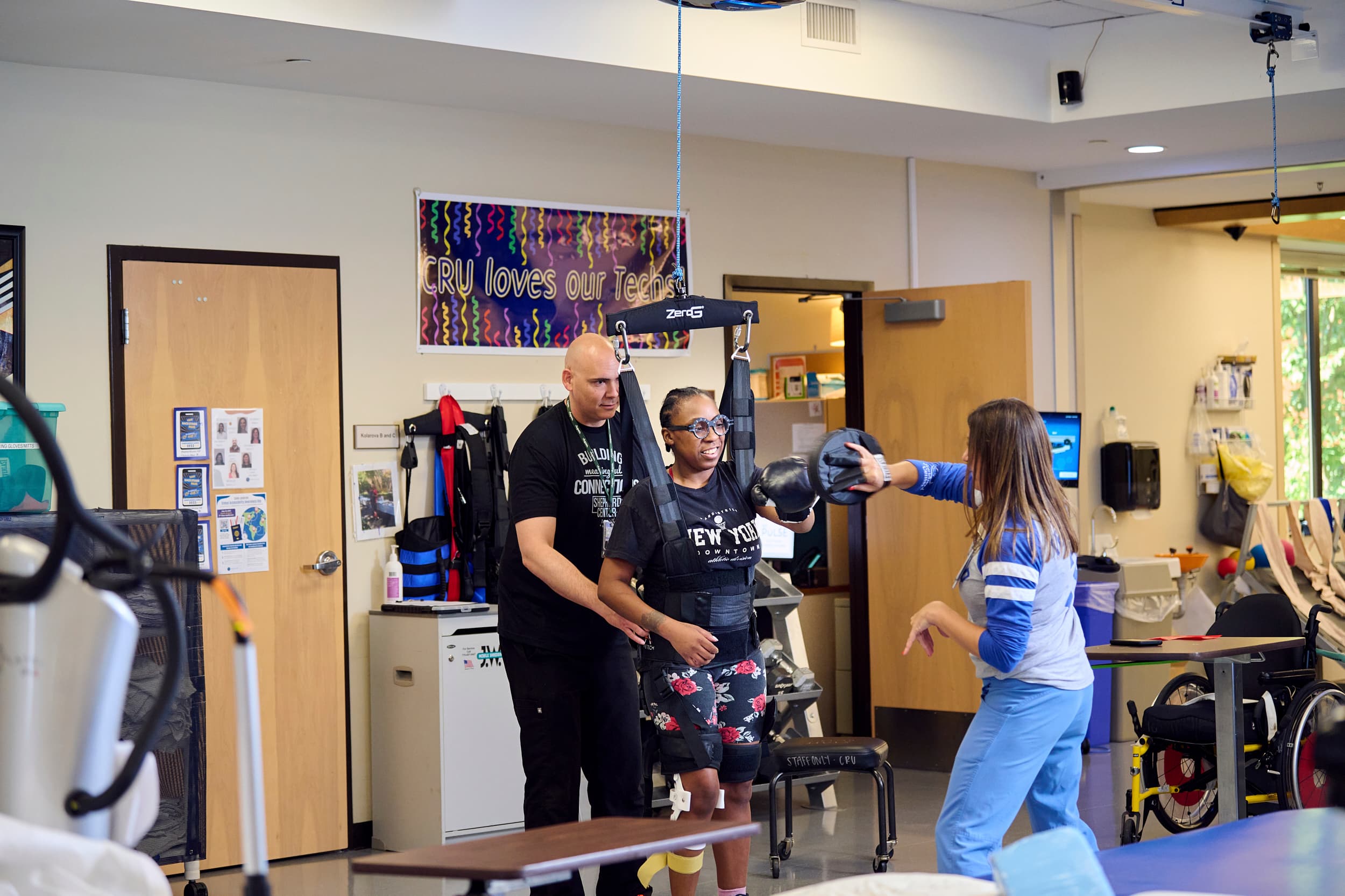 A young woman undergoes physical therapy, supported by two therapists. One therapist helps her stand using a harness system, while the other assists with arm exercises. The room has motivational posters and various rehabilitation equipment.