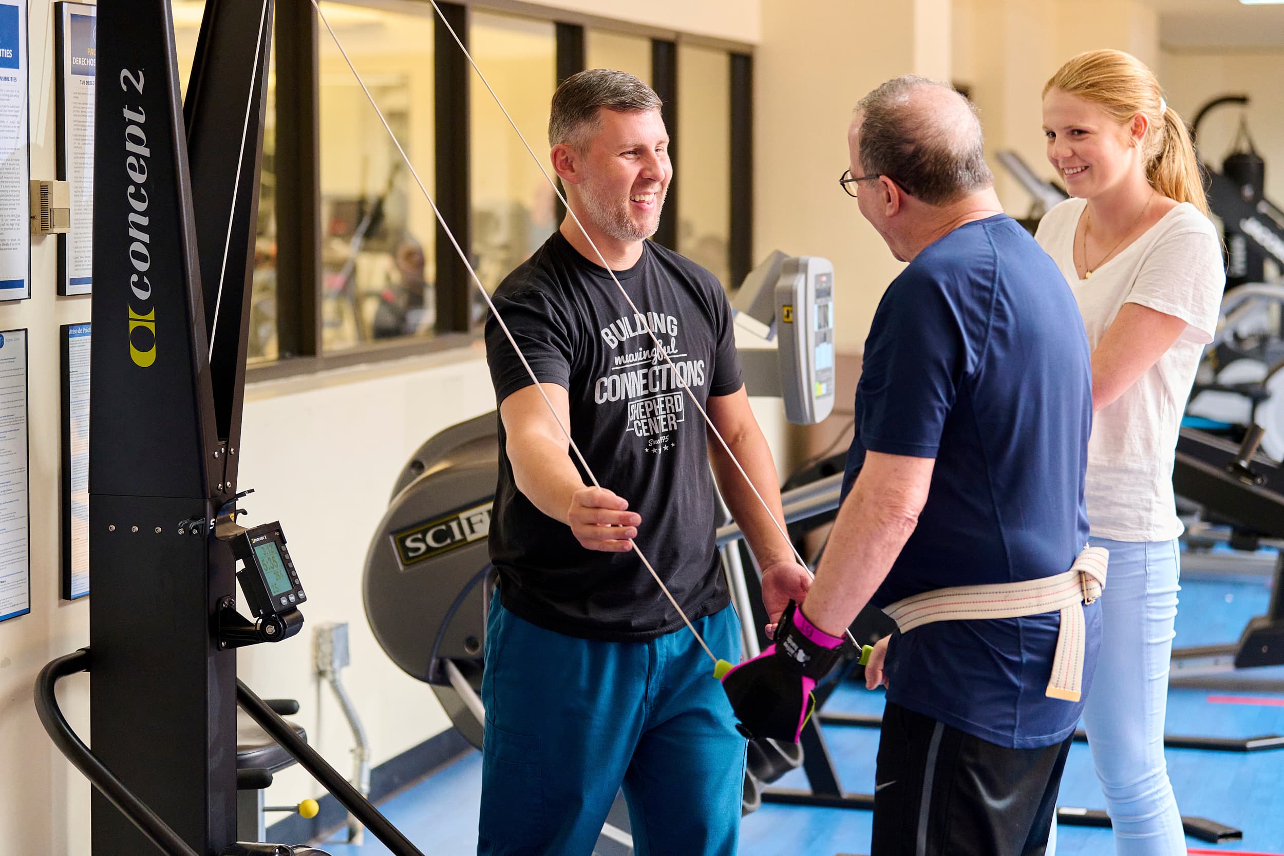 Three people in a gym: a man using exercise equipment with assistance from another man and a woman. They are engaged in a friendly interaction. Exercise machines and windows are visible in the background.