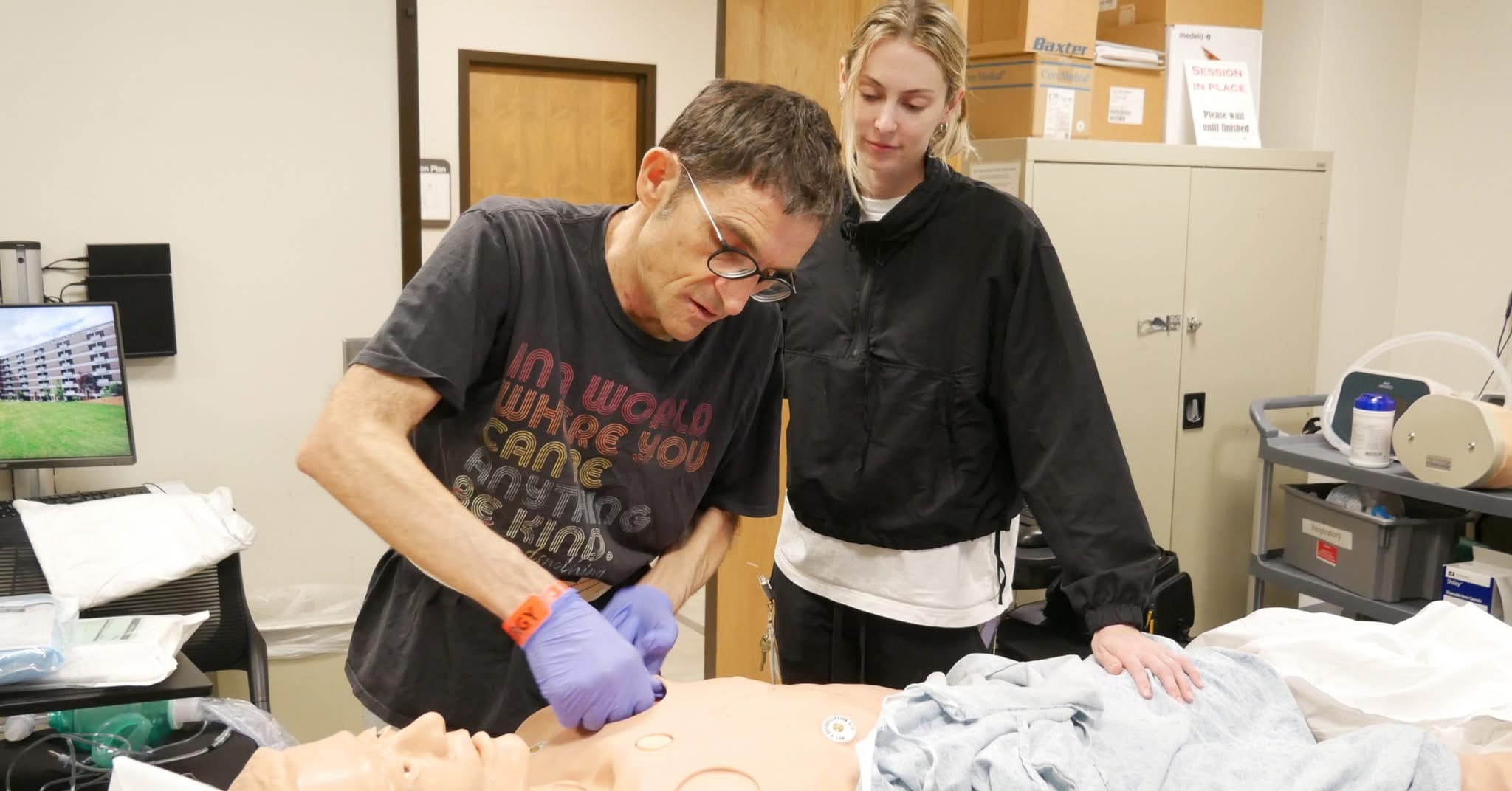 Two people practice a medical procedure on a mannequin in a clinical setting. One person, wearing gloves, demonstrates, while the other observes attentively. Medical equipment and supplies are visible in the background.