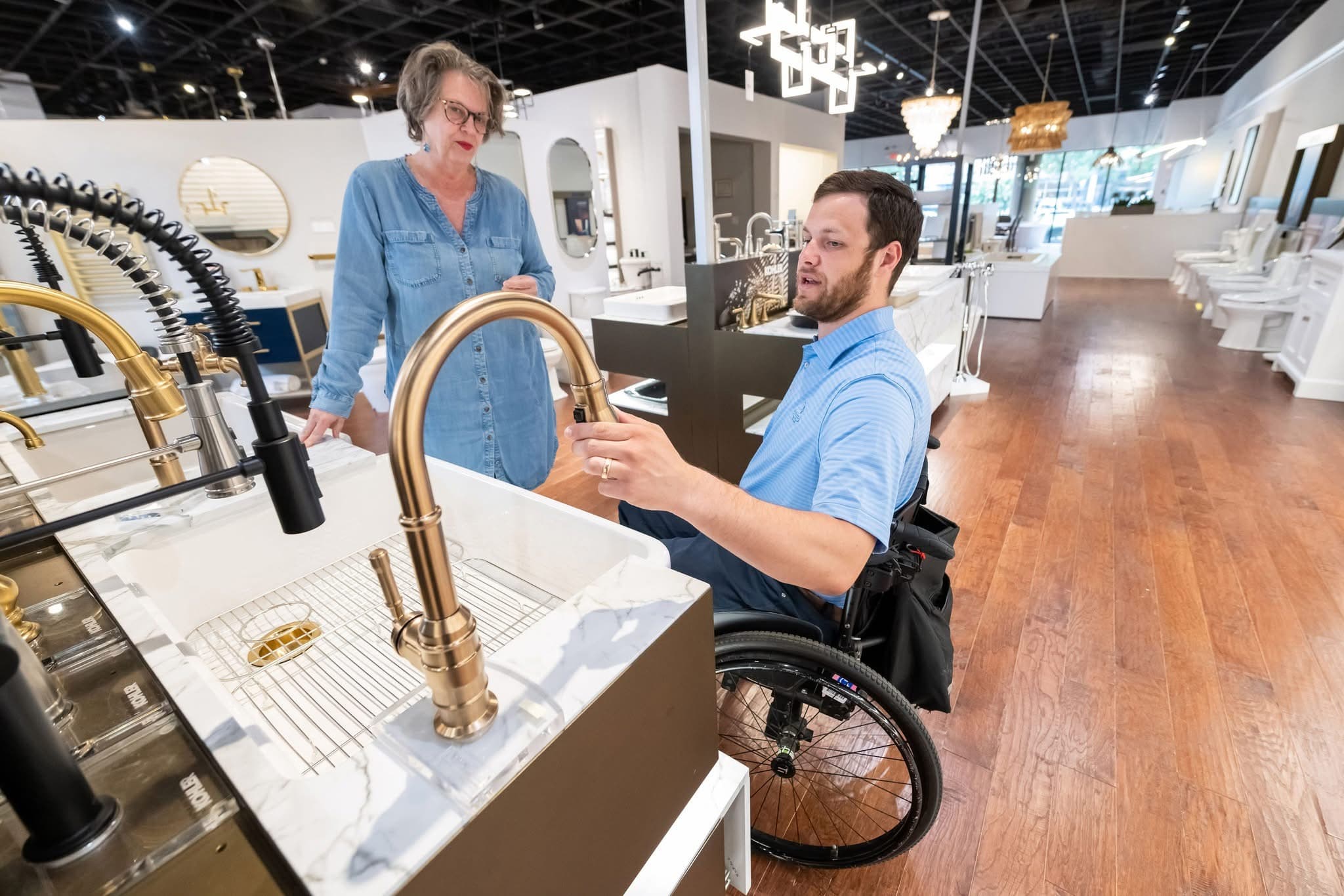 A man in a wheelchair examines a gold faucet display in a showroom, while a woman standing beside him engages in conversation. The showroom features a variety of modern fixtures and wooden flooring.