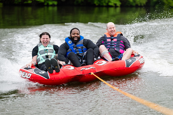 Three people in life jackets joyfully ride on a red inflatable tube being pulled across a lake. Water splashes behind them, with lush green trees in the background.