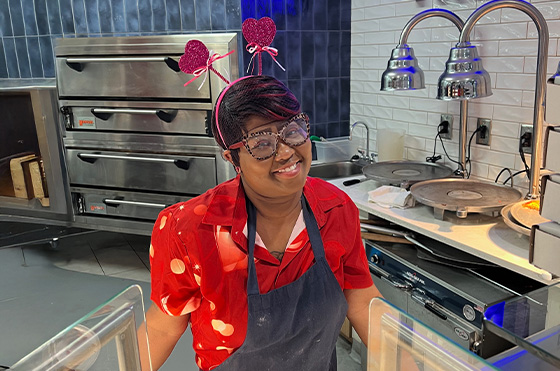 A person wearing a festive headband with sparkling hearts and large glasses stands behind a food counter, smiling. They are dressed in a red shirt with white polka dots and a dark apron. Pizza ovens and warming lights are visible in the background.