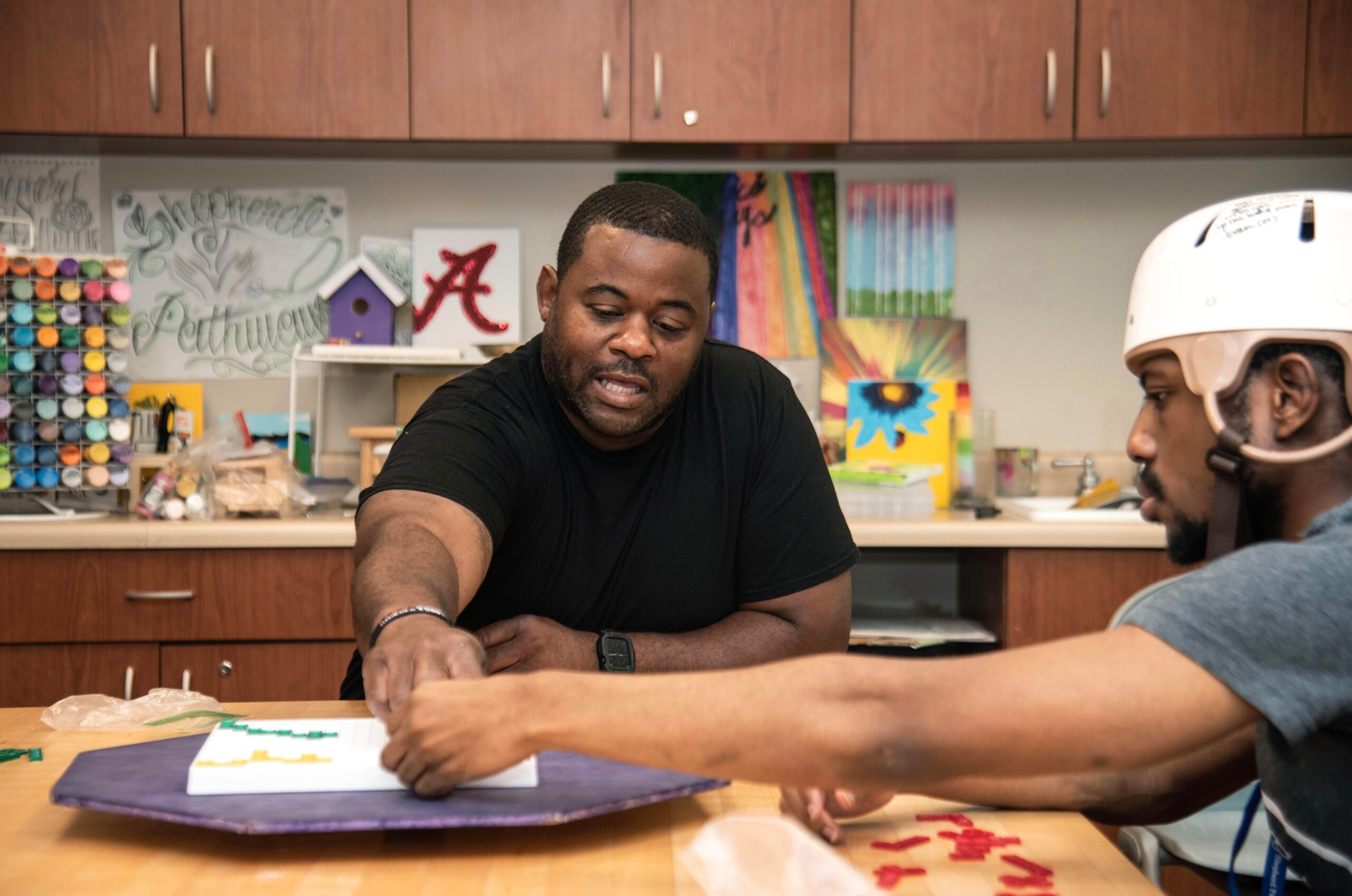 A teacher and a student are seated at a table engaged in an art project. The student is wearing a protective helmet. The classroom is filled with art supplies and colorful paintings. Both individuals are focused on the task at hand.