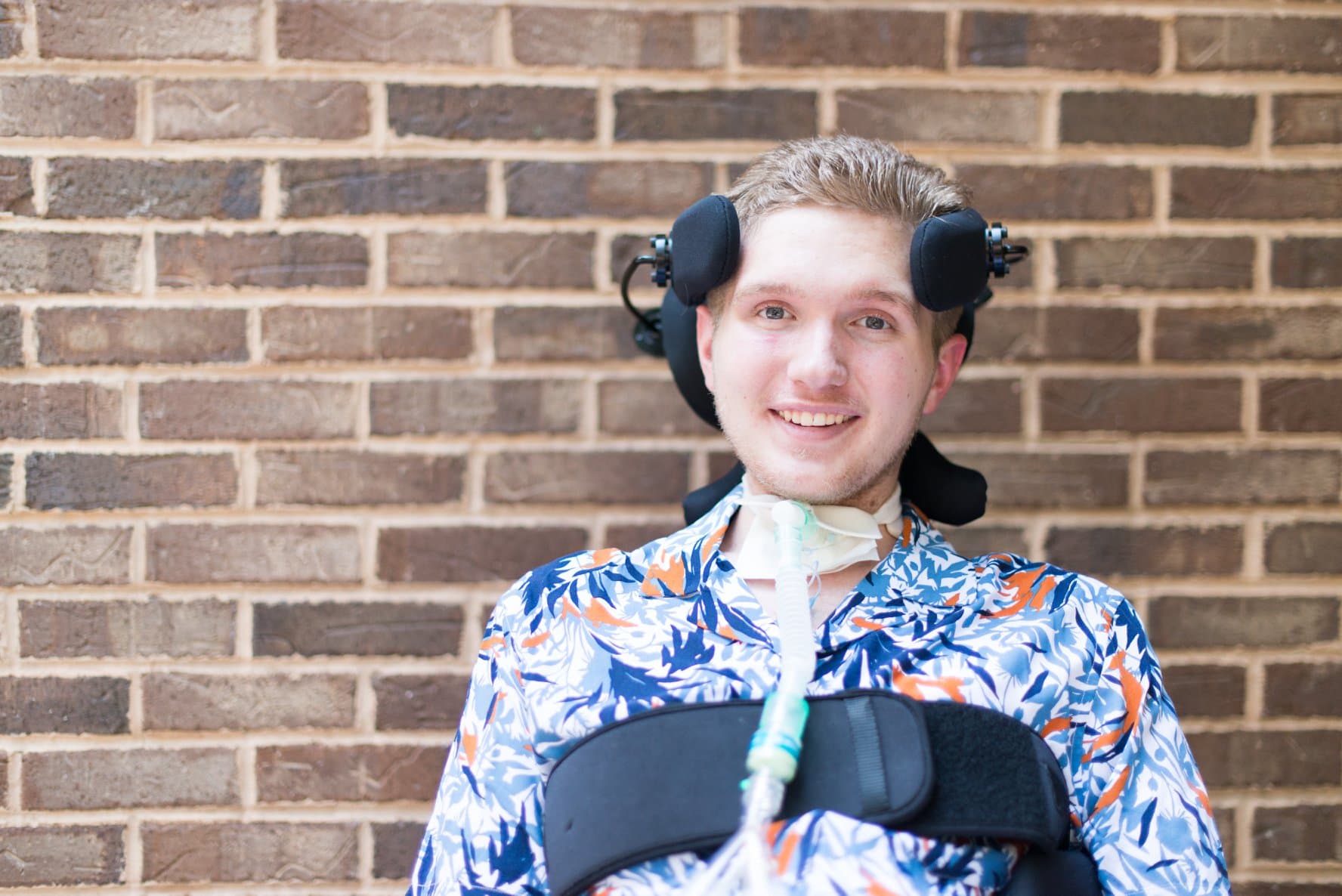 A young man with light brown hair smiles while sitting against a brick wall. He wears a colorful floral shirt and a supportive harness, with respiratory equipment visible around his neck.