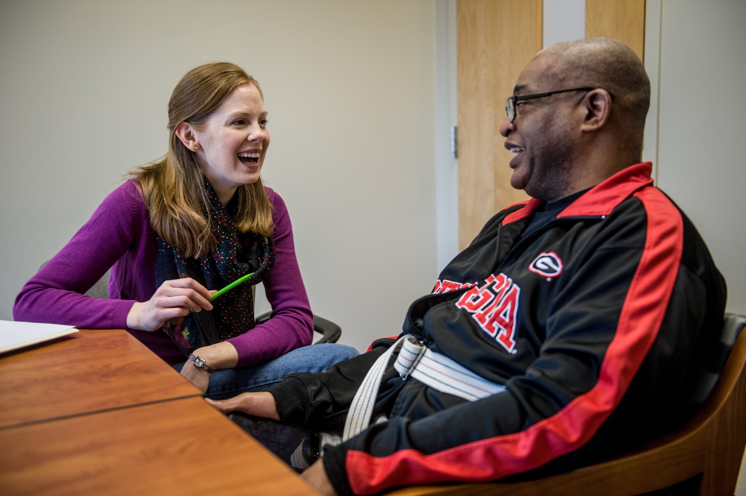 A woman and a man are sitting at a table, engaged in a friendly conversation. The woman is holding a pen and smiling, while the man, wearing a Georgia sports jacket, is also smiling. They're in a room with a wooden door in the background.