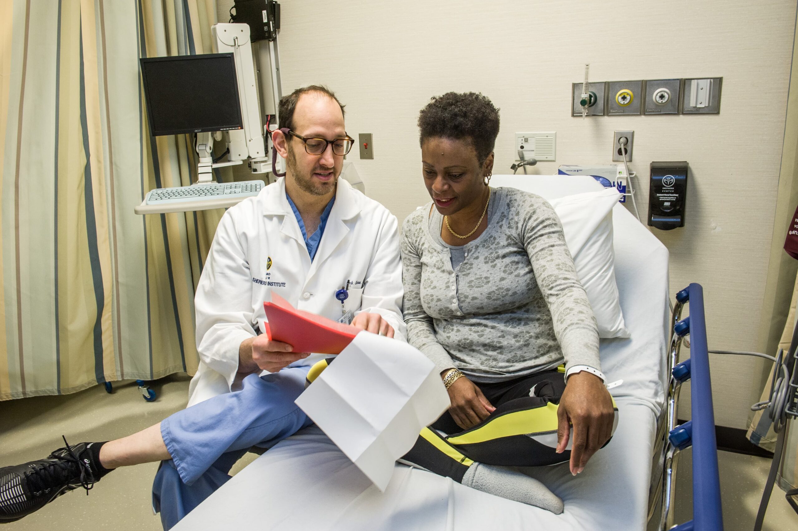 A doctor, clad in a white coat, is explaining paperwork to a patient suffering from chronic pain. The patient, seated and dressed in casual clothes, listens intently in the hospital room filled with medical equipment and a bed.