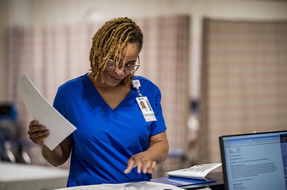 A healthcare professional in blue scrubs stands at a desk, holding papers and smiling. A computer screen and an open binder are visible in the foreground. The background is softly blurred, suggesting a clinical setting.