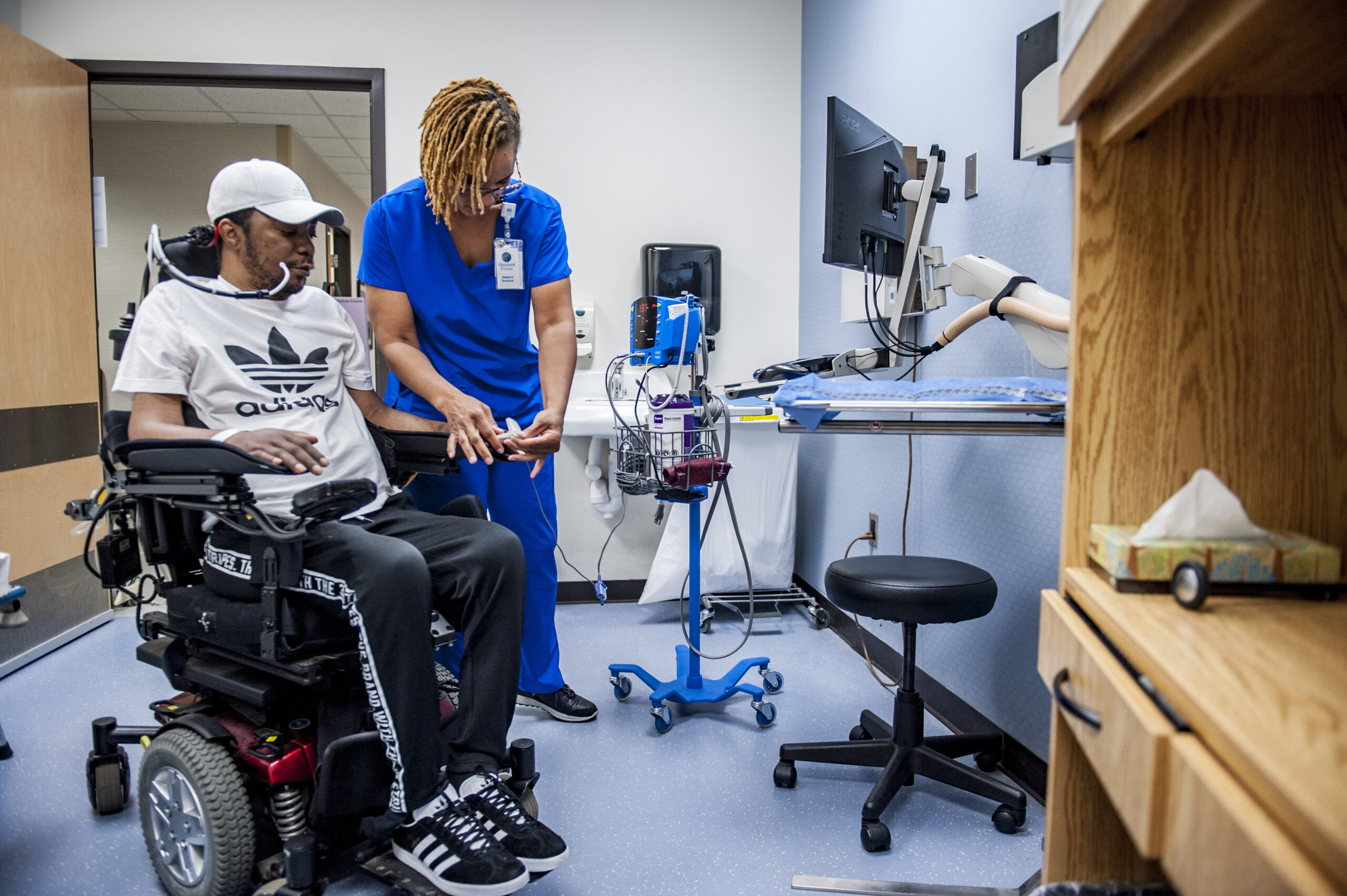 A healthcare professional in blue scrubs assists a person in a wheelchair inside a medical office, where neurourology treatment is available. The room contains medical equipment, a monitor, and shelves with supplies.