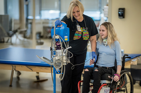 A healthcare professional assists a smiling young woman in a wheelchair with a medical device. The setting appears to be a clinical environment, with a table visible in the background. Both individuals are engaged and appear to be in good spirits.