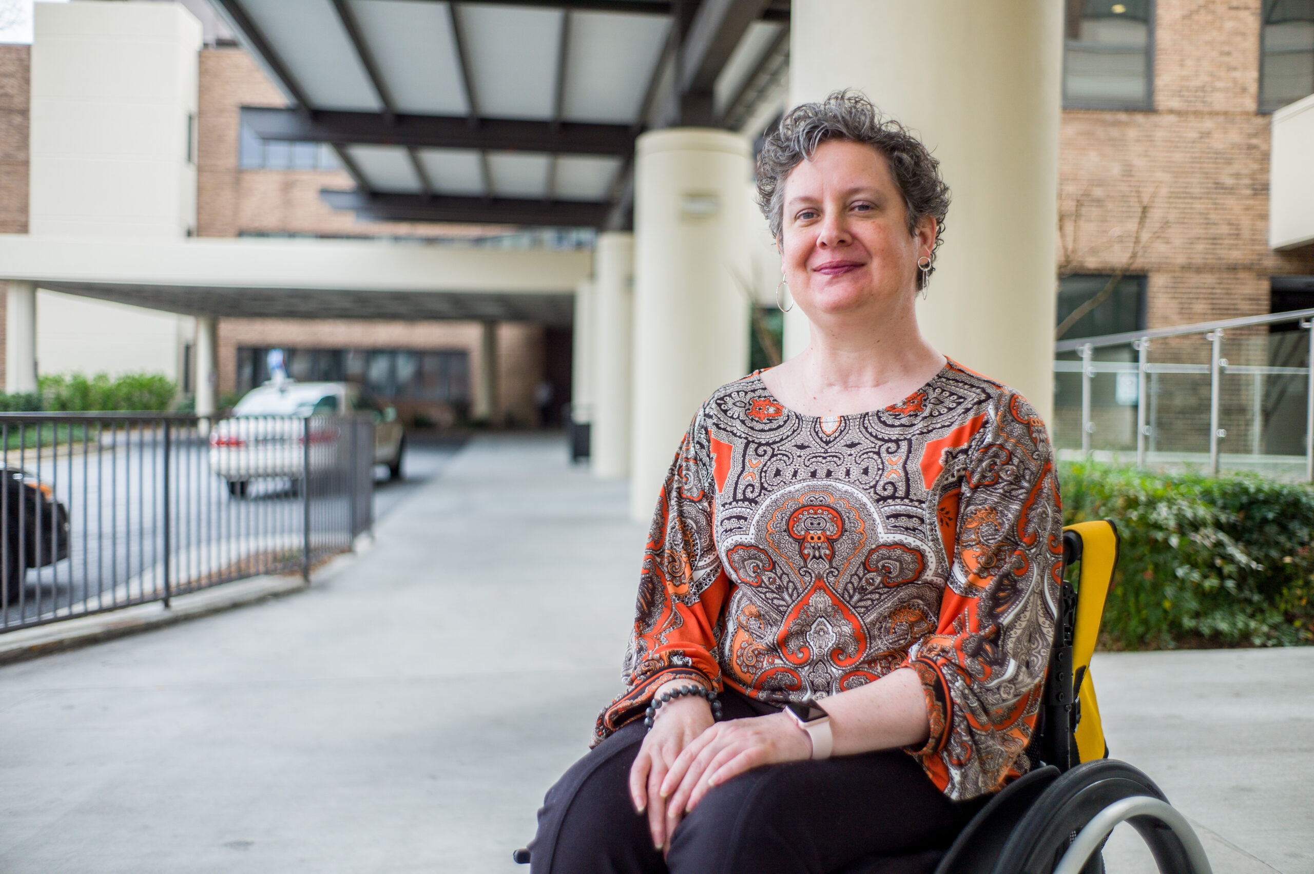 A person with short, curly hair sits in a wheelchair outdoors near a building, perhaps pondering 