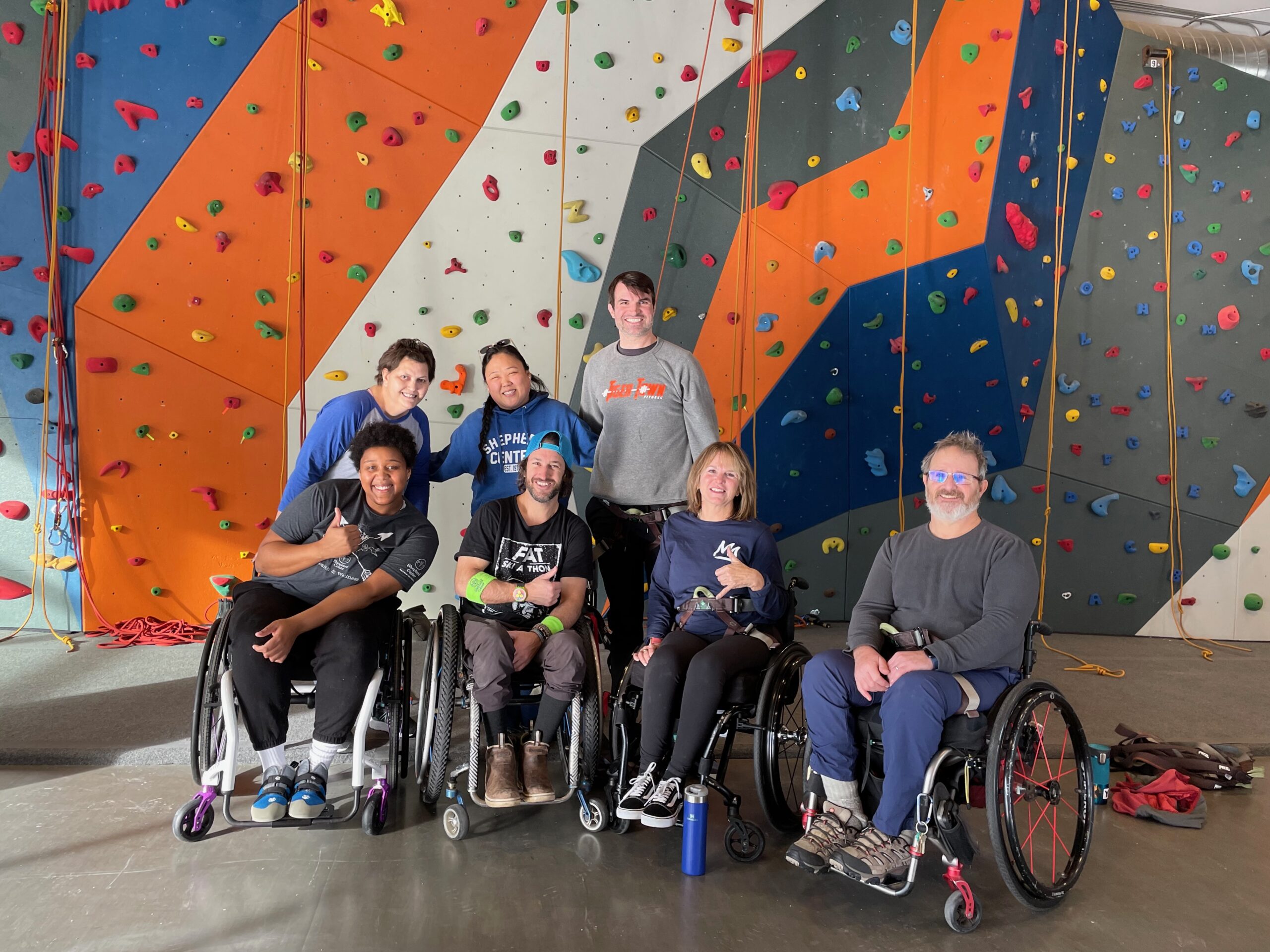 A person in a blue outfit, with climbing gear, is smiling while being suspended in a climbing gym. The background shows a climbing wall with colorful holds and several people.