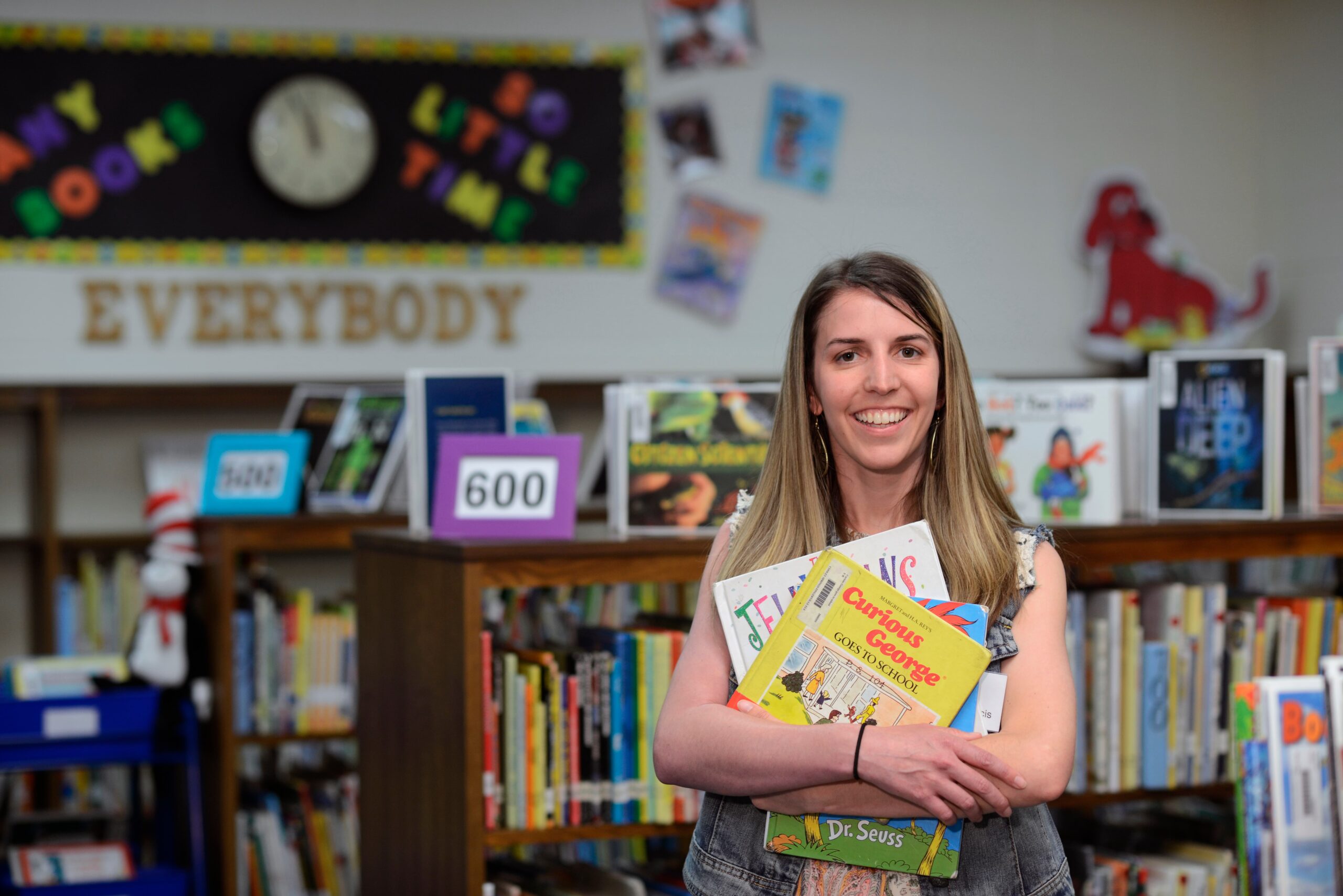 Former patient, Amanda Francis, holding a stack of children's books in her arms while smiling while standing in a school library.